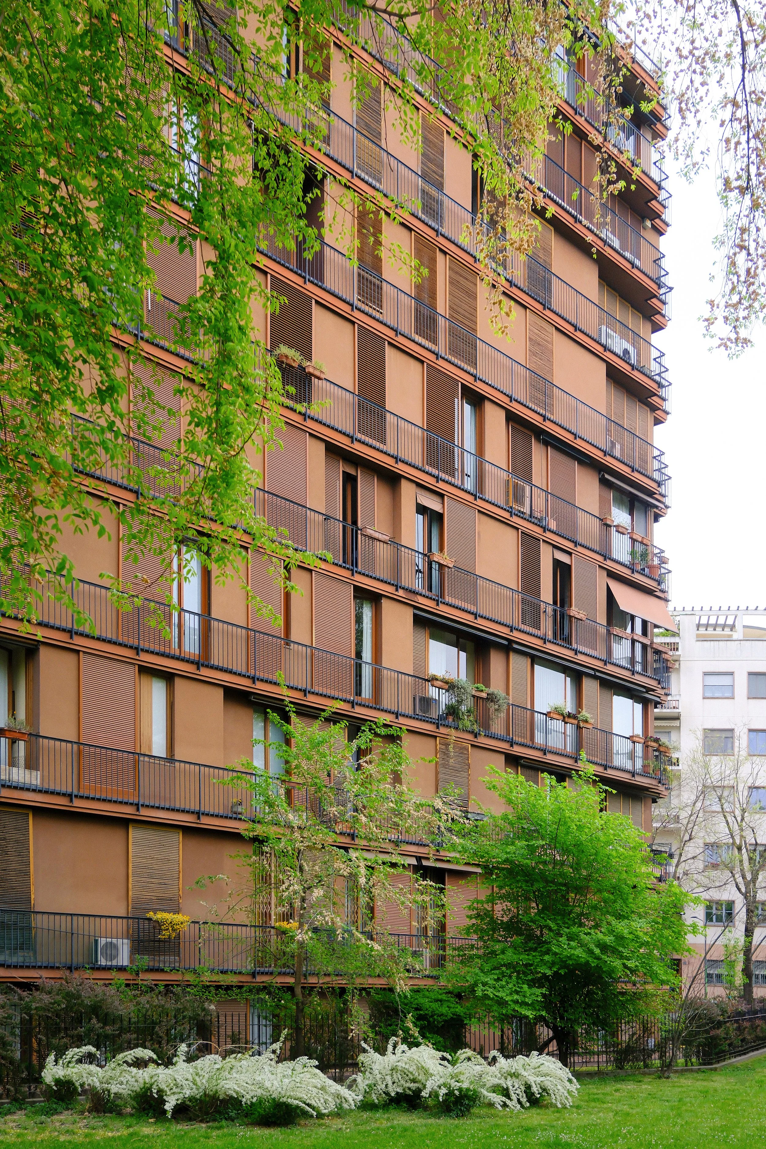 A tall residential building with multiple balconies and brown window shutters, surrounded by green trees and bushes, with some white flowering plants in the foreground.