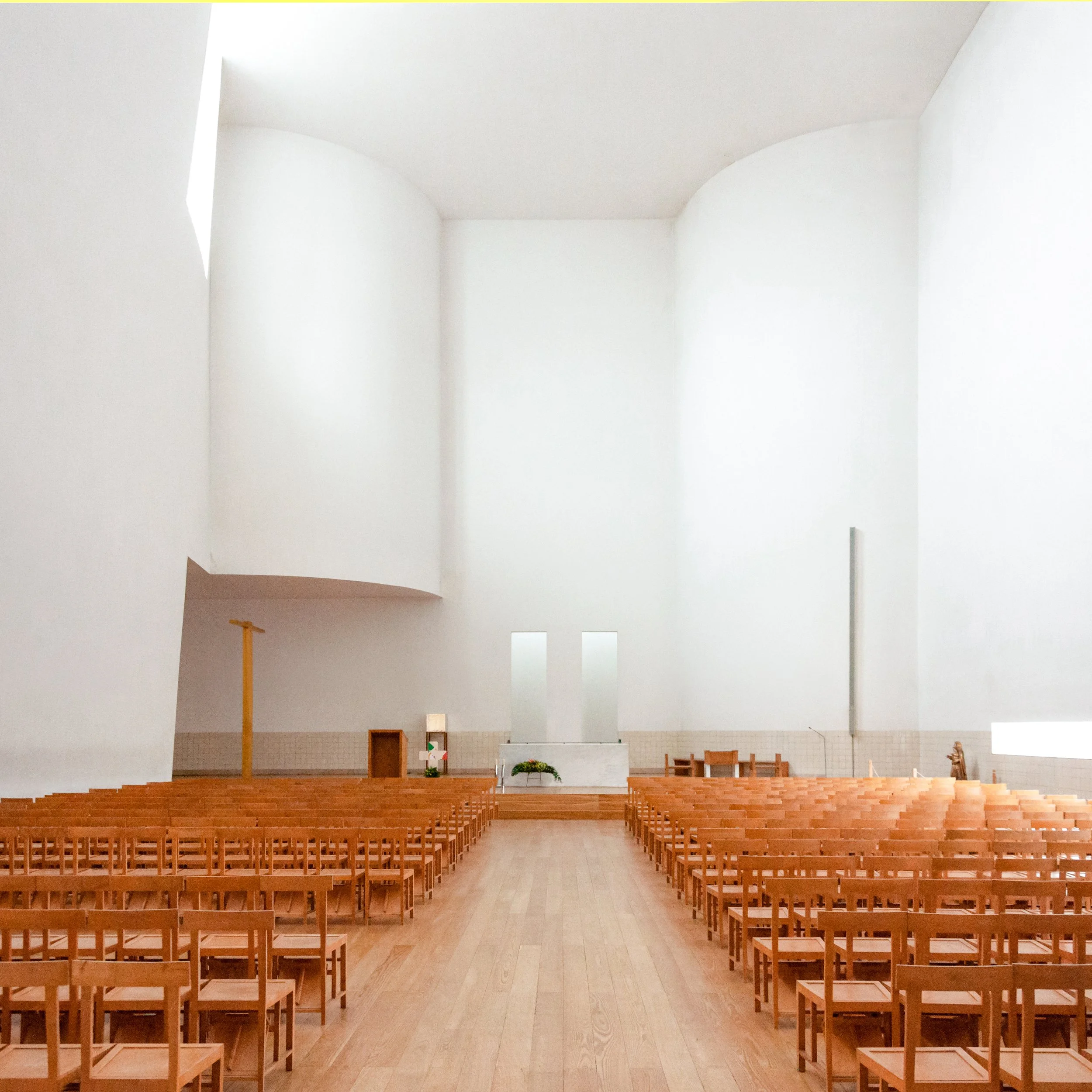 Inside a minimalist church with white walls and a high ceiling, rows of wooden chairs face an altar with flowers. The space is bright with natural light.