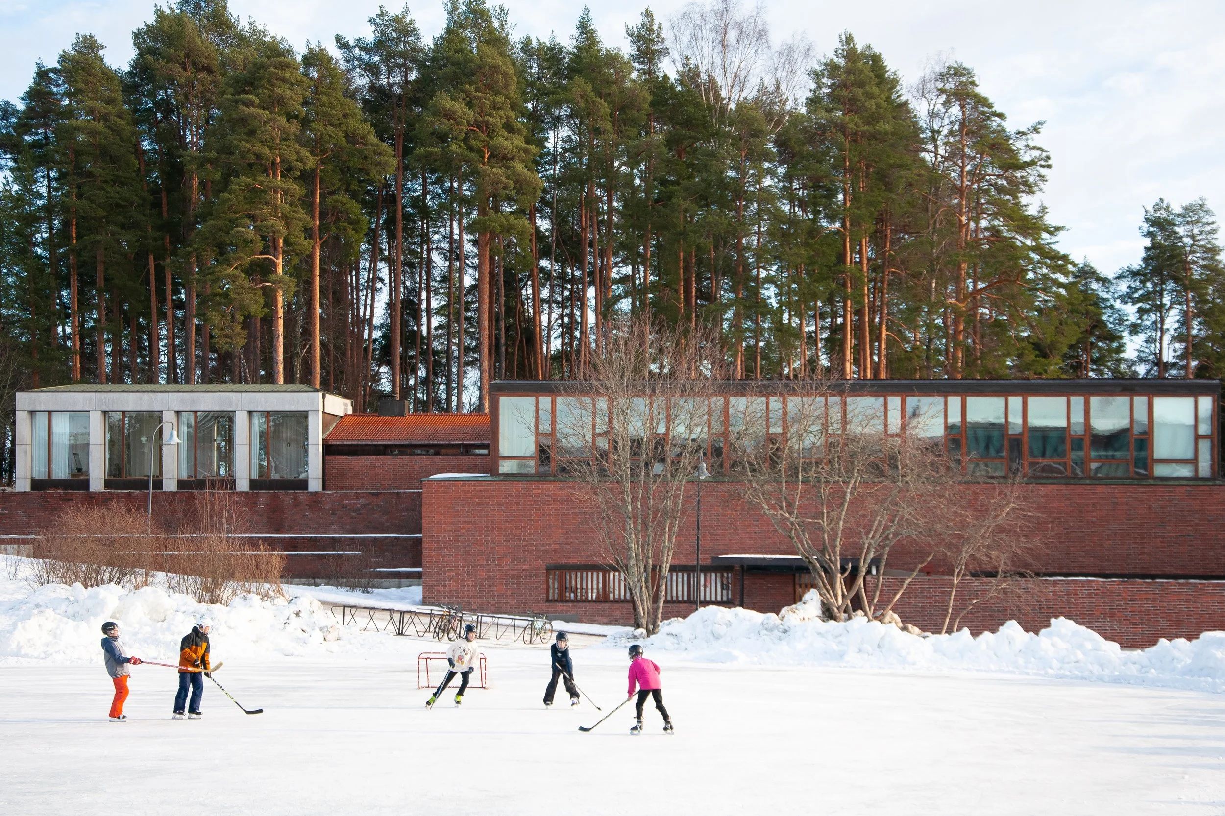 Children playing ice hockey on an outdoor rink surrounded by snow with trees and modern brick building in the background.