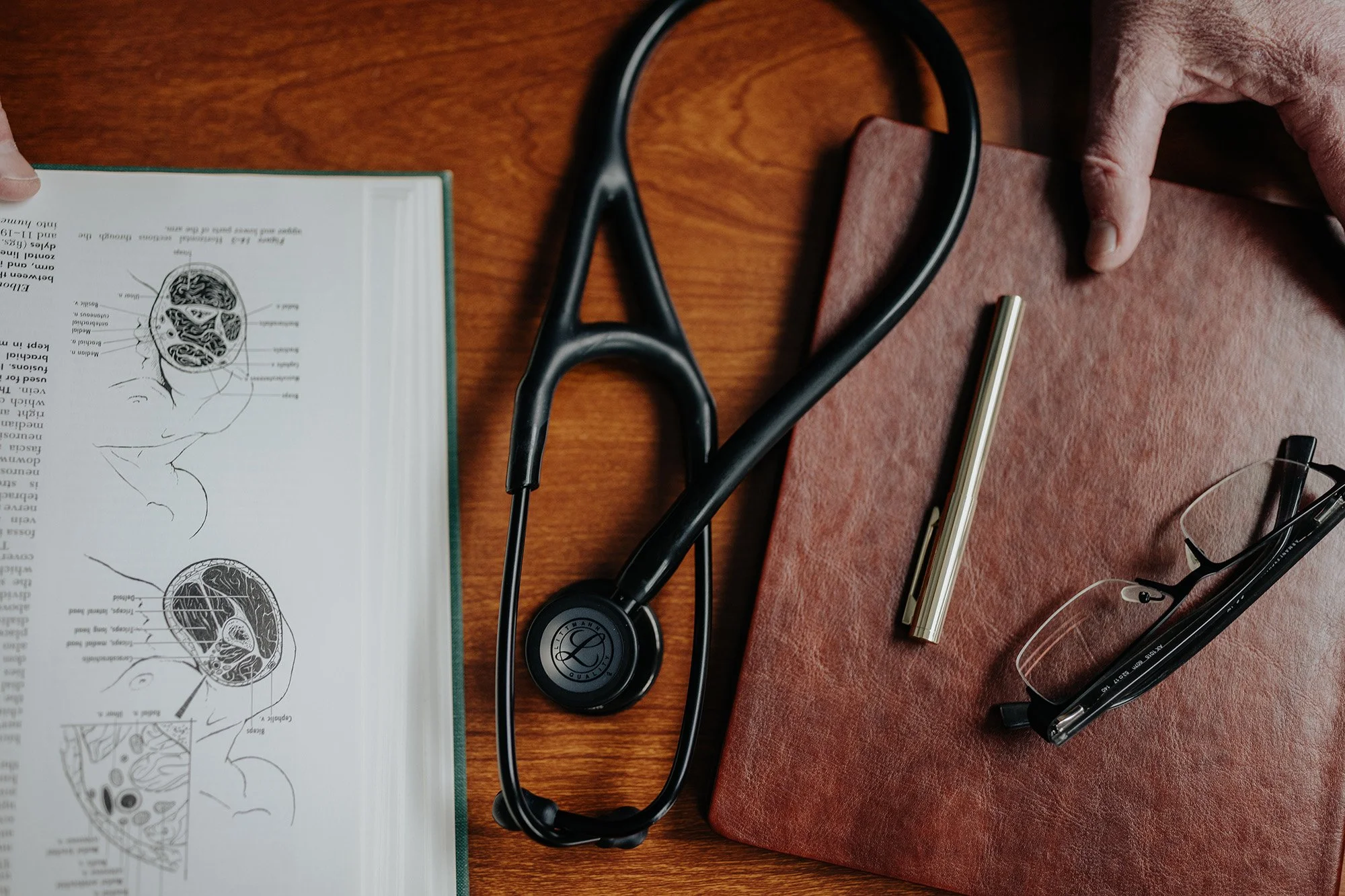 A stethoscope, a red notebook, a pen, safety glasses, and a medical diagram of the human brain on a wooden desk.