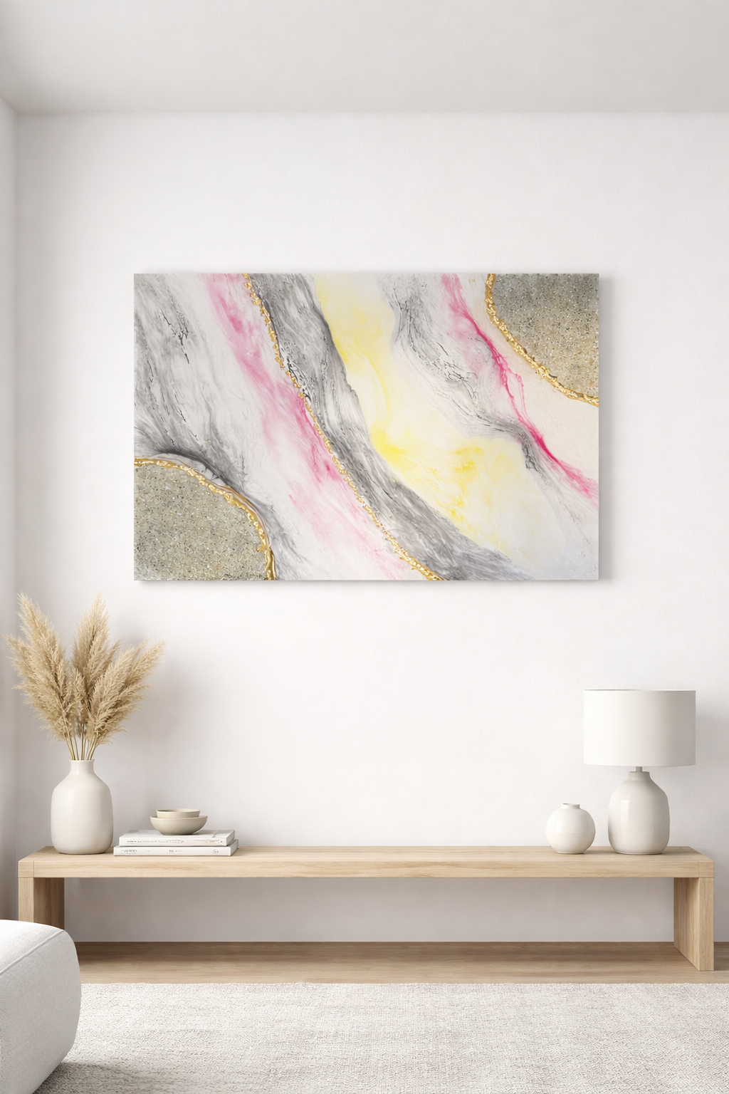 Abstract wall art with pink, yellow, black, gray, and gold accents hanging above a wooden console table with a white vase of dried grasses, a stack of books, and white decor items in a minimalist, modern room.