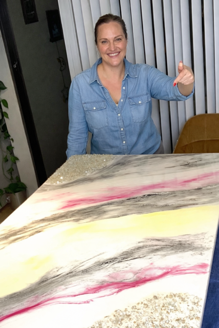 A smiling woman in a denim shirt giving a thumbs-up, standing behind a colorful abstract art piece on a table.