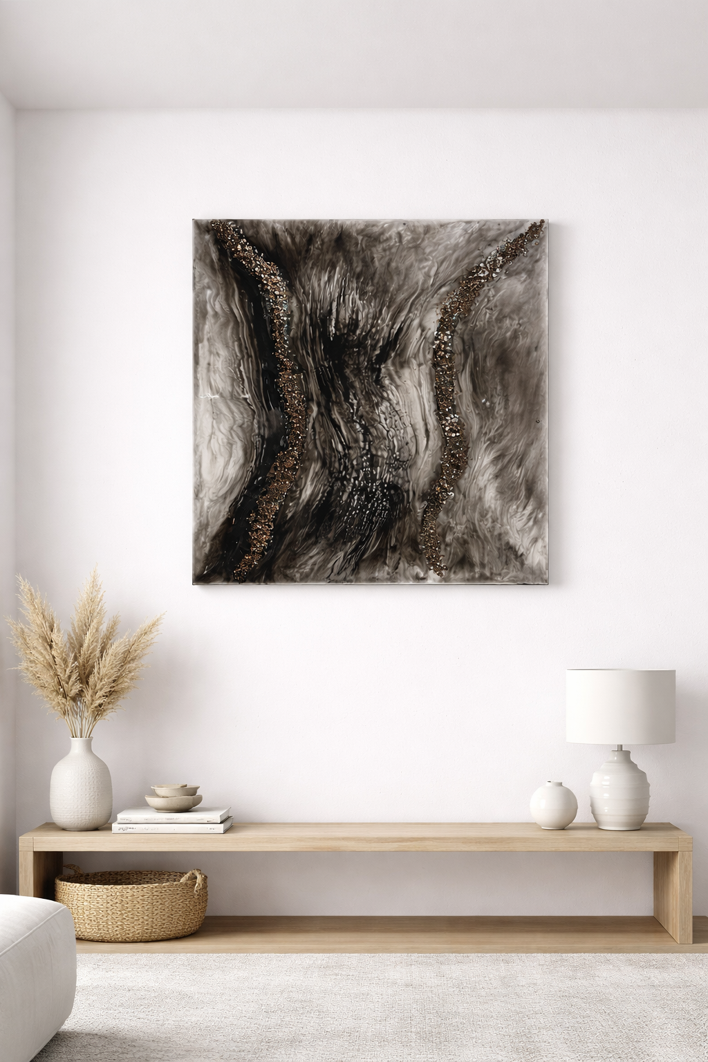 Abstract black, gray, gold, and white painting hung on a white wall above a wooden console table with decor including a vase with dried pampas grass, small bowls, a lamp, and decorative vases.
