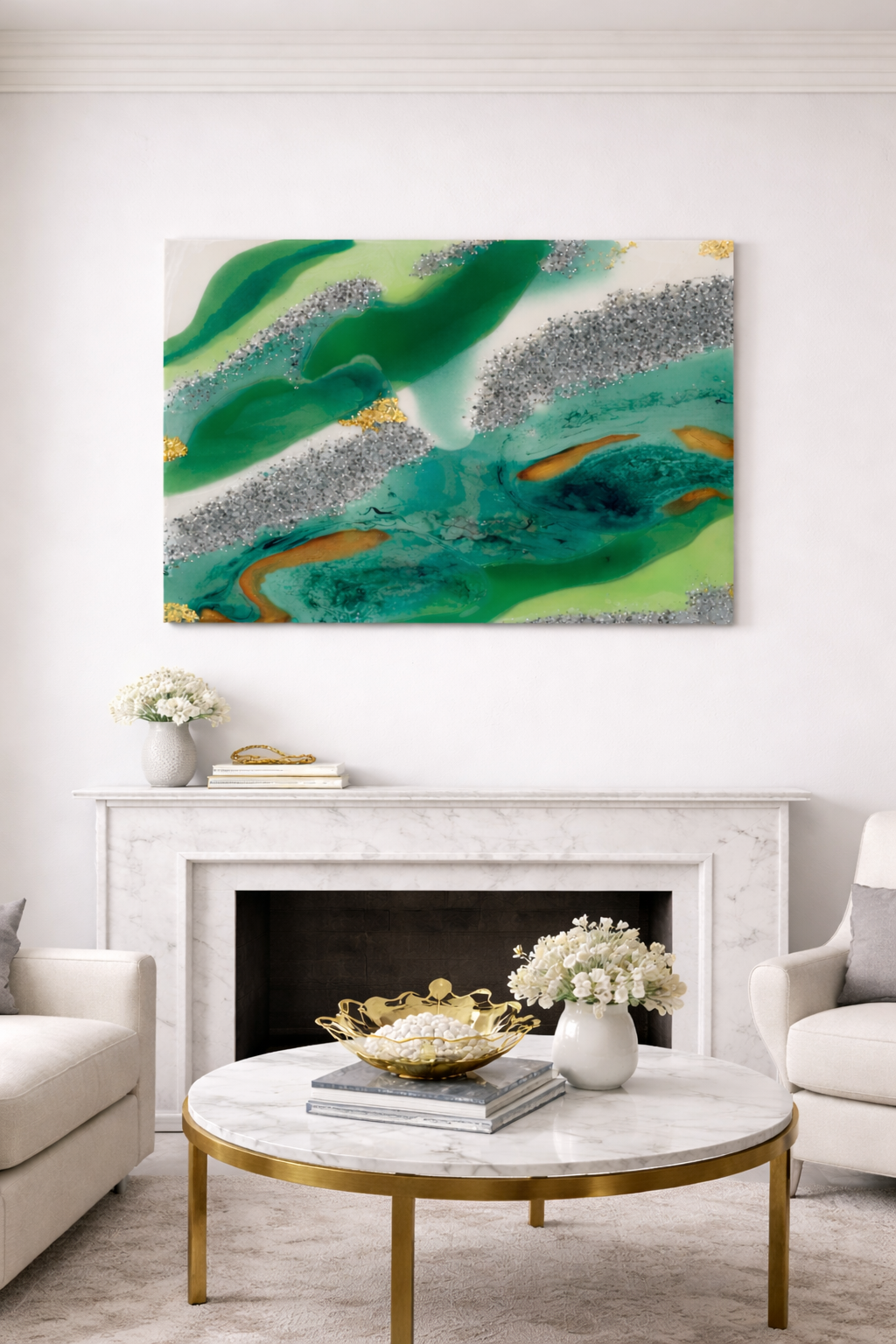 An abstract painting with green, blue, gray, and gold colors hanging above a white marble fireplace in a modern living room. The fireplace mantel holds a white vase with flowers and some books. There is a round marble coffee table with a gold rim decorated with a gold bowl, a white vase with flowers, and a stack of books. Two white armchairs with gray pillows are on either side of the table.