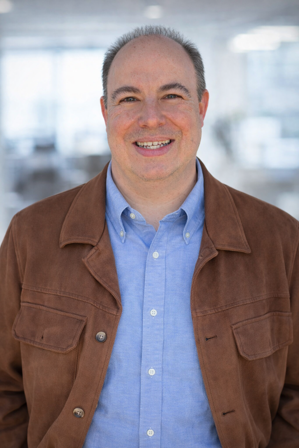 A smiling man with short gray hair, wearing a light blue shirt and a brown jacket, standing in a bright indoor setting.
