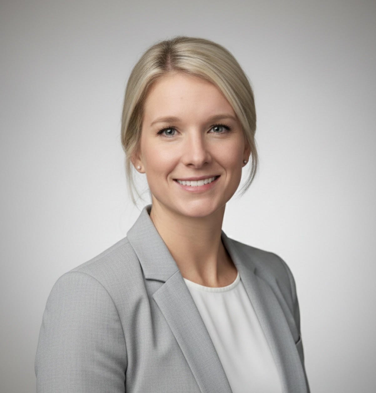 A professional woman with blonde hair and blue eyes, smiling, wearing a light gray blazer and white top against a plain gray background.