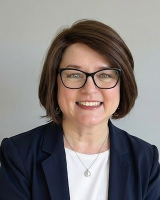 A woman with short brown hair, black glasses, smiling, wearing a dark blazer and a white top, against a plain light background.