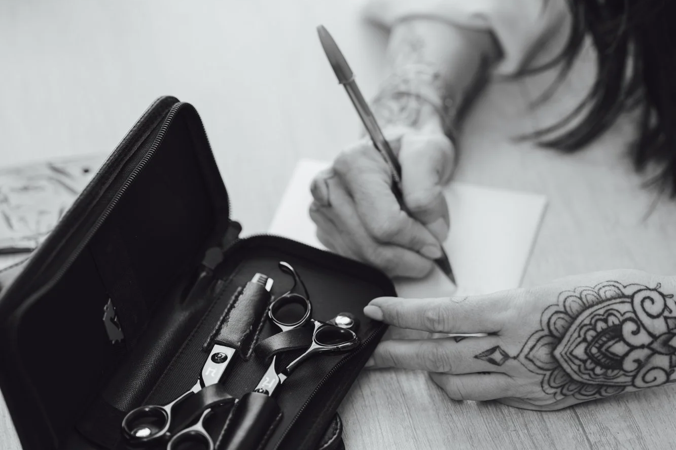 A person with intricate tattoo designs on their hands is writing on a piece of paper with a pen. A case containing tattoo scissors, a comb, and other tattoo tools is open on the table.