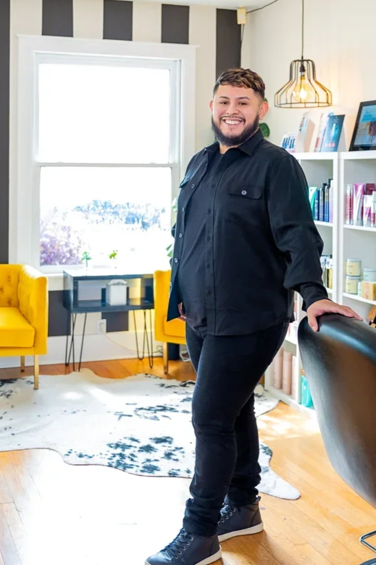 A man with a beard and short hair, wearing a black jacket and pants, standing in a modern living room with bright yellow chairs, white bookshelf, and a large window with a snow-covered landscape outside.