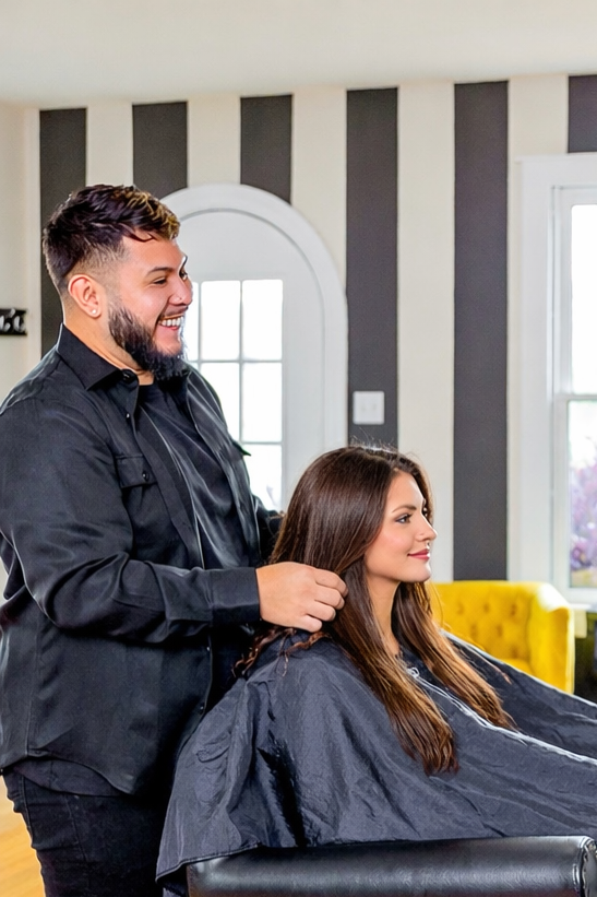 A hairstylist is cutting a woman's hair in a bright, modern salon with black-and-white striped walls and large windows.