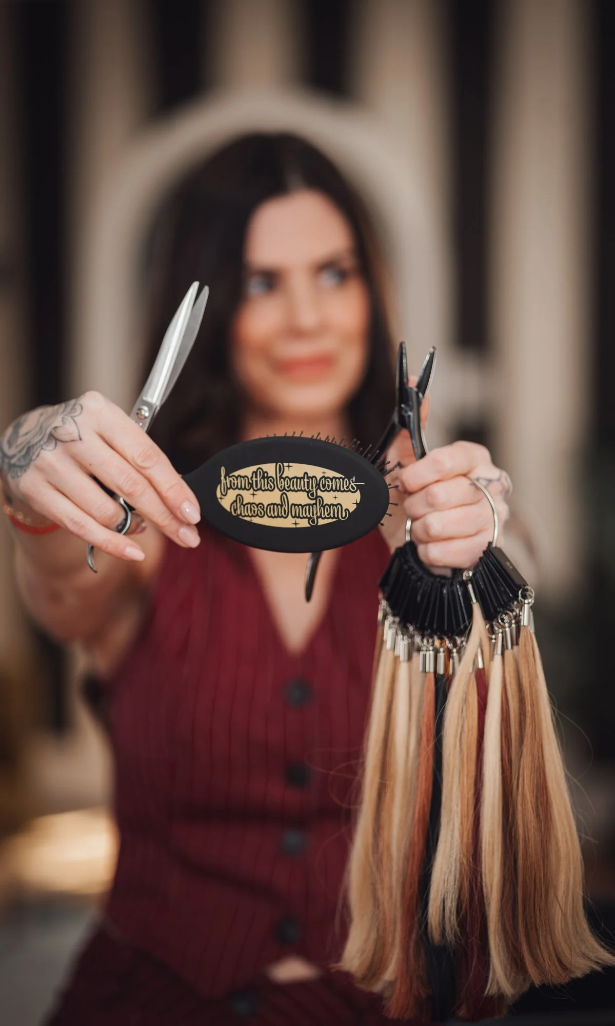 A woman with dark hair holding hair color swatches and hairdressing scissors in a salon. The hair swatches are attached to keyrings, and one keyring has a black sign with gold lettering that reads 'from this beauty come chaos and mayhem.'