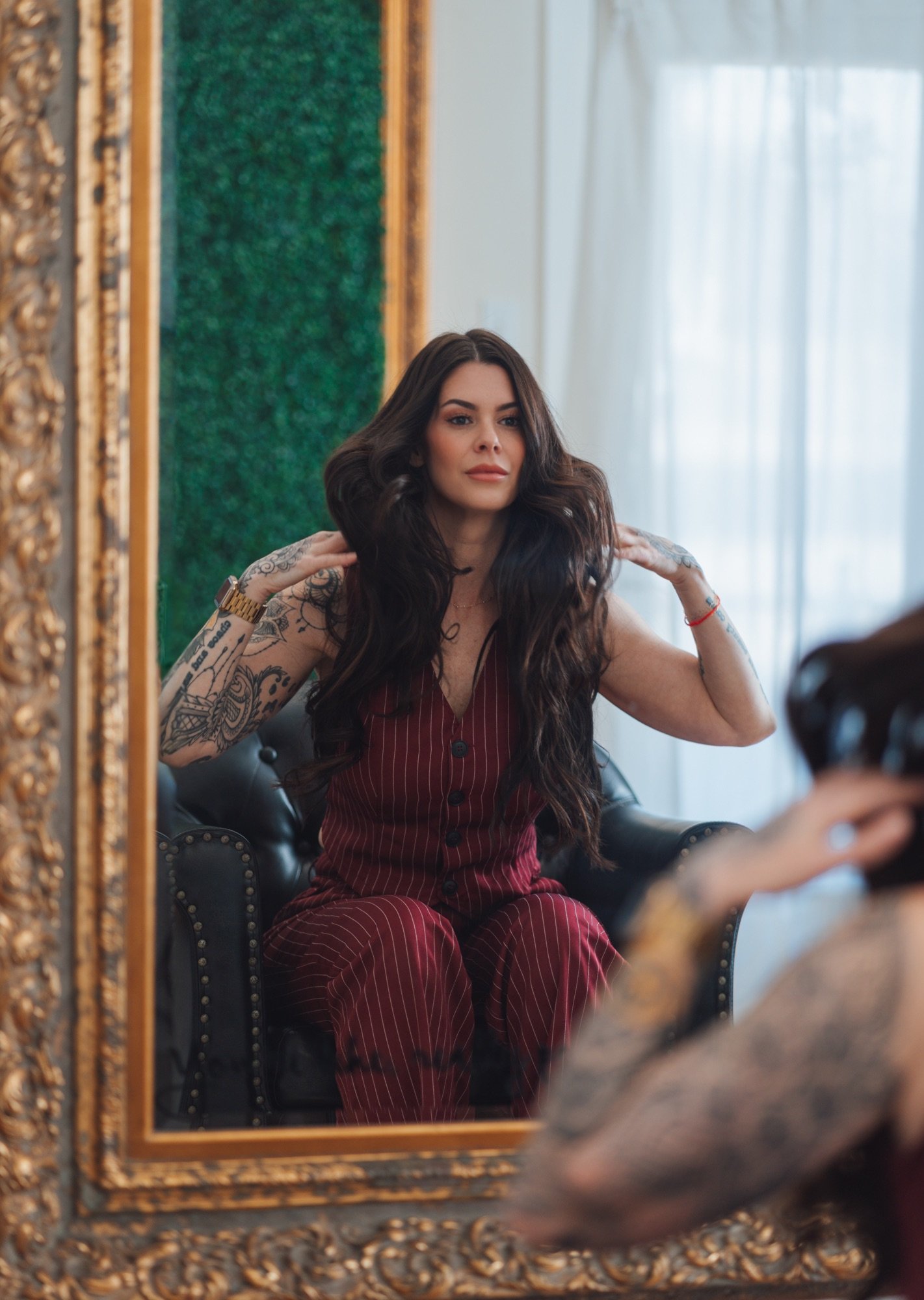 Woman with long dark hair and tattoos, sitting on a black leather chair, adjusting her hair, reflected in an ornate gold-framed mirror, in a room with a green wall and sheer white curtains.