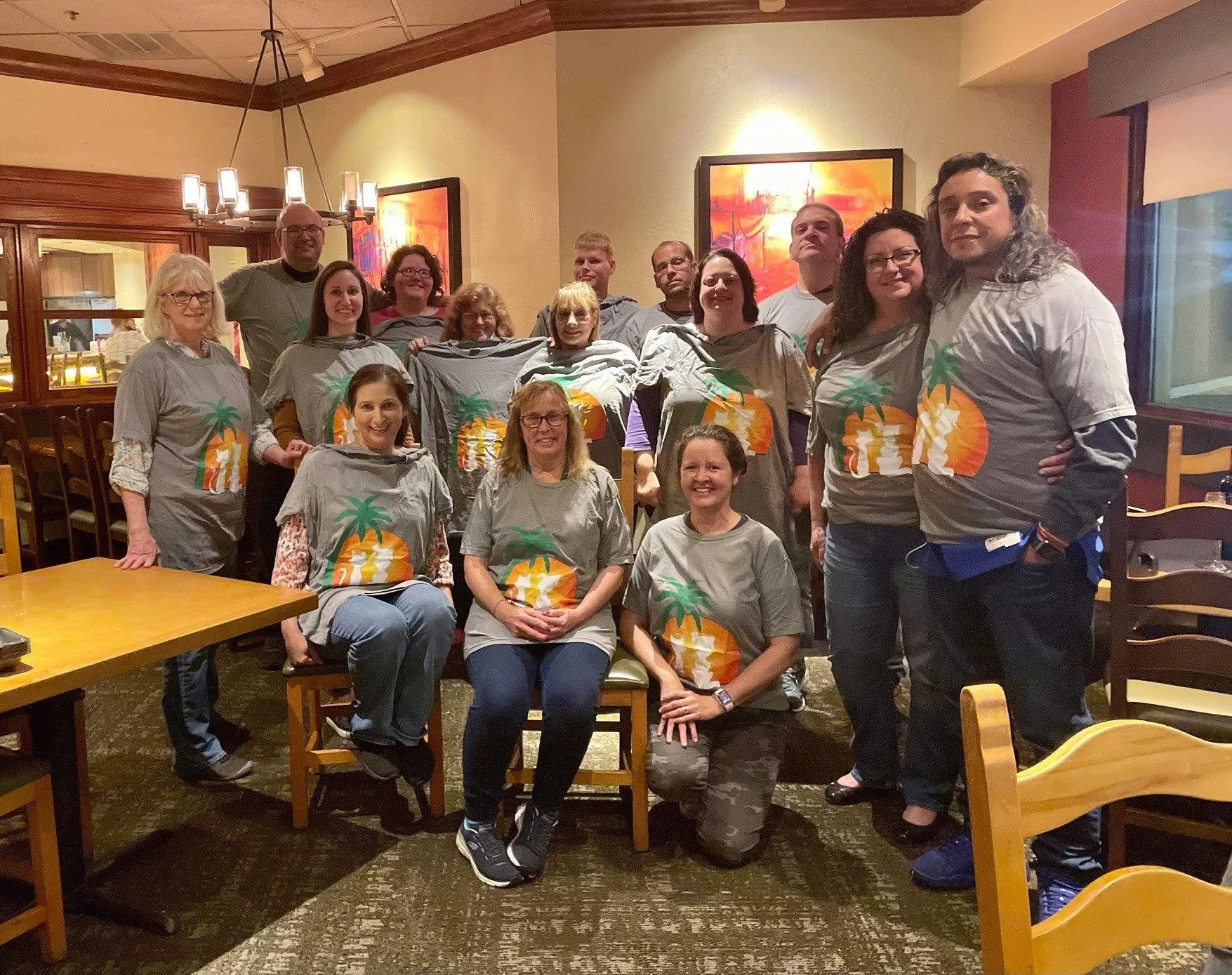 Group of people posing together in a restaurant, some wearing gray t-shirts with a tropical island graphic, in front of warm-colored artwork on the wall.