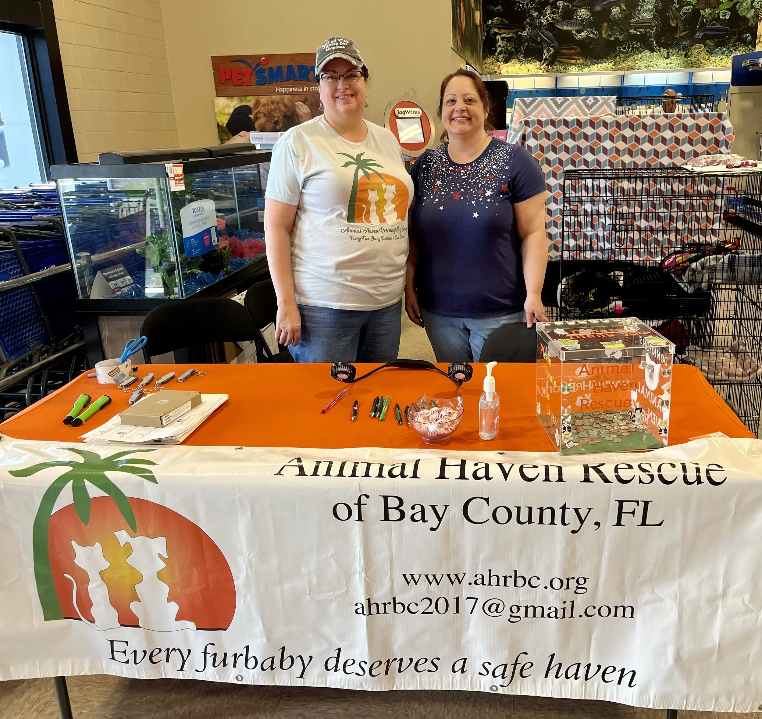 Two women standing behind a table at an Animal Haven Rescue event inside a store. The table has a banner with the rescue's name, website, and email, featuring a logo of two cats and a dog. Items on the table include pens, a fidget spinner, a donation box, hand sanitizer, and paperwork. Behind them, shelves with pet supplies and an aquarium are visible.
