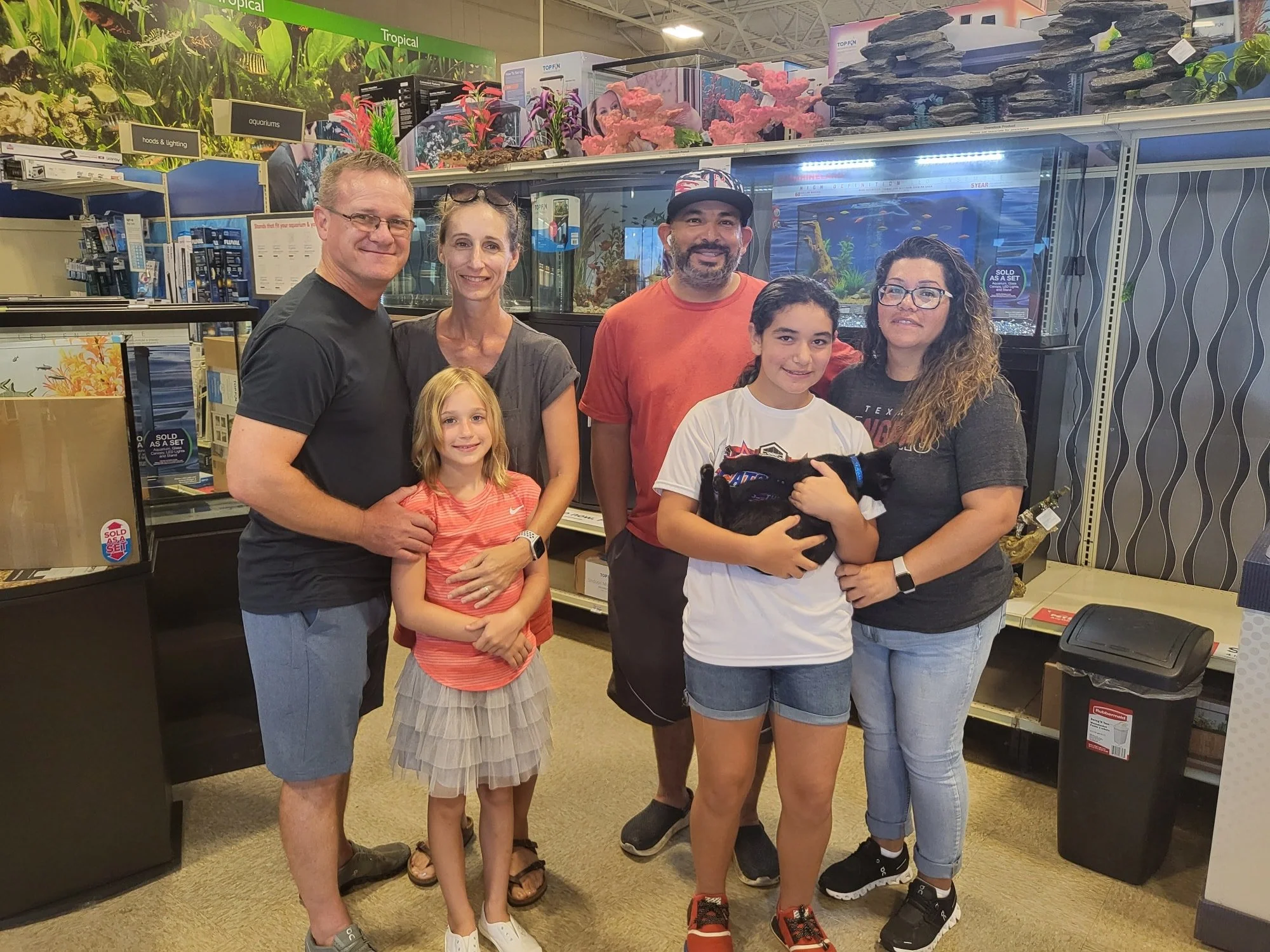 Group of six people, including adults and children, posing together inside a pet or aquarium store, holding a black kitten. Behind them are aquariums with water and aquatic plants.
