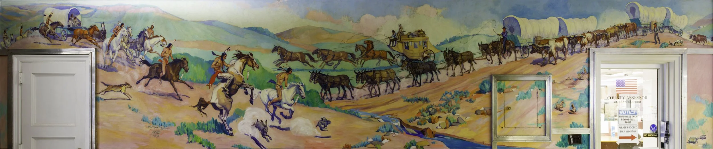 Colorful mural depicting a historic wagon train of covered wagons and horses with Native American figures in a desert landscape with mountains, shrubs, and a stream