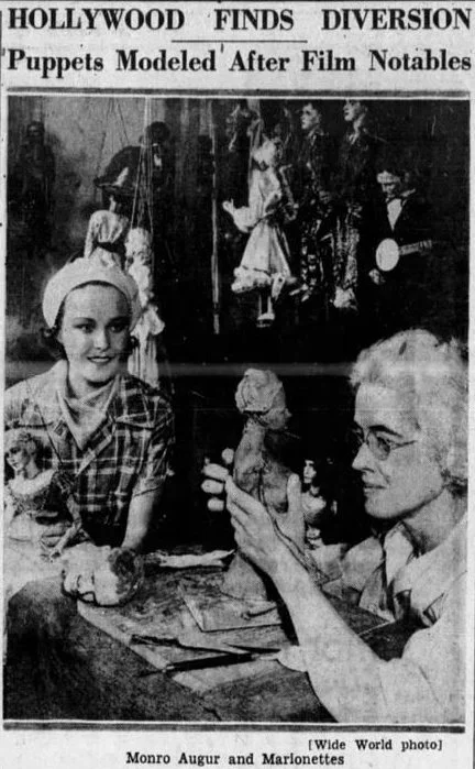 A black-and-white newspaper photo showing two women in the foreground, one younger with a headscarf and the other older with glasses, sitting at a table. The older woman is holding a puppet. In the background, there is a stage with puppeteers and puppets, and a headline reading 'Hollywood Finds Diversion Puppets Modeled After Film Notables'. The caption indicates the women are Monroe Augur and Marlonettes.
