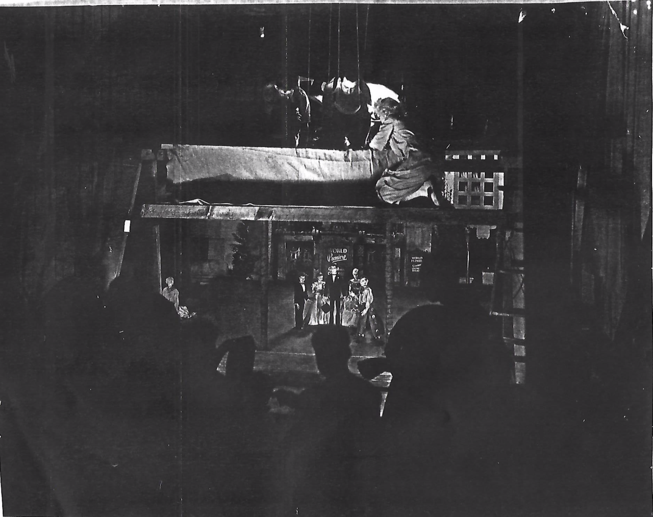 Black and white photograph of a puppet show on a stage with a building facade backdrop, and spectators in the foreground watching.