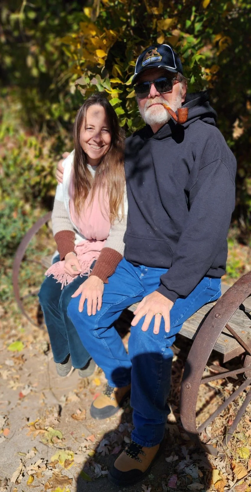 A smiling woman with long brown hair, wearing a pink dress and a beige cardigan with brown sleeves, sitting next to an older man with white hair and a beard, wearing a black hoodie, blue jeans, sunglasses, a cap, and smoking a pipe. They are sitting on a wooden bench outdoors surrounded by autumn leaves and green foliage.
