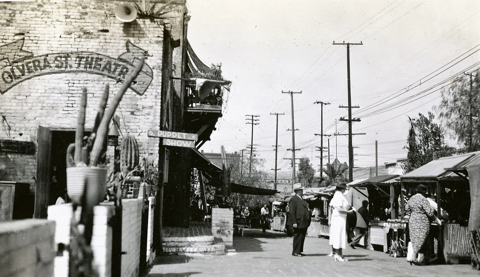 Historical black and white photo of a street market scene with people shopping at outdoor stalls, power lines overhead, and a brick building with signage for a puppet show, cactus plants, and a few trees in the background.