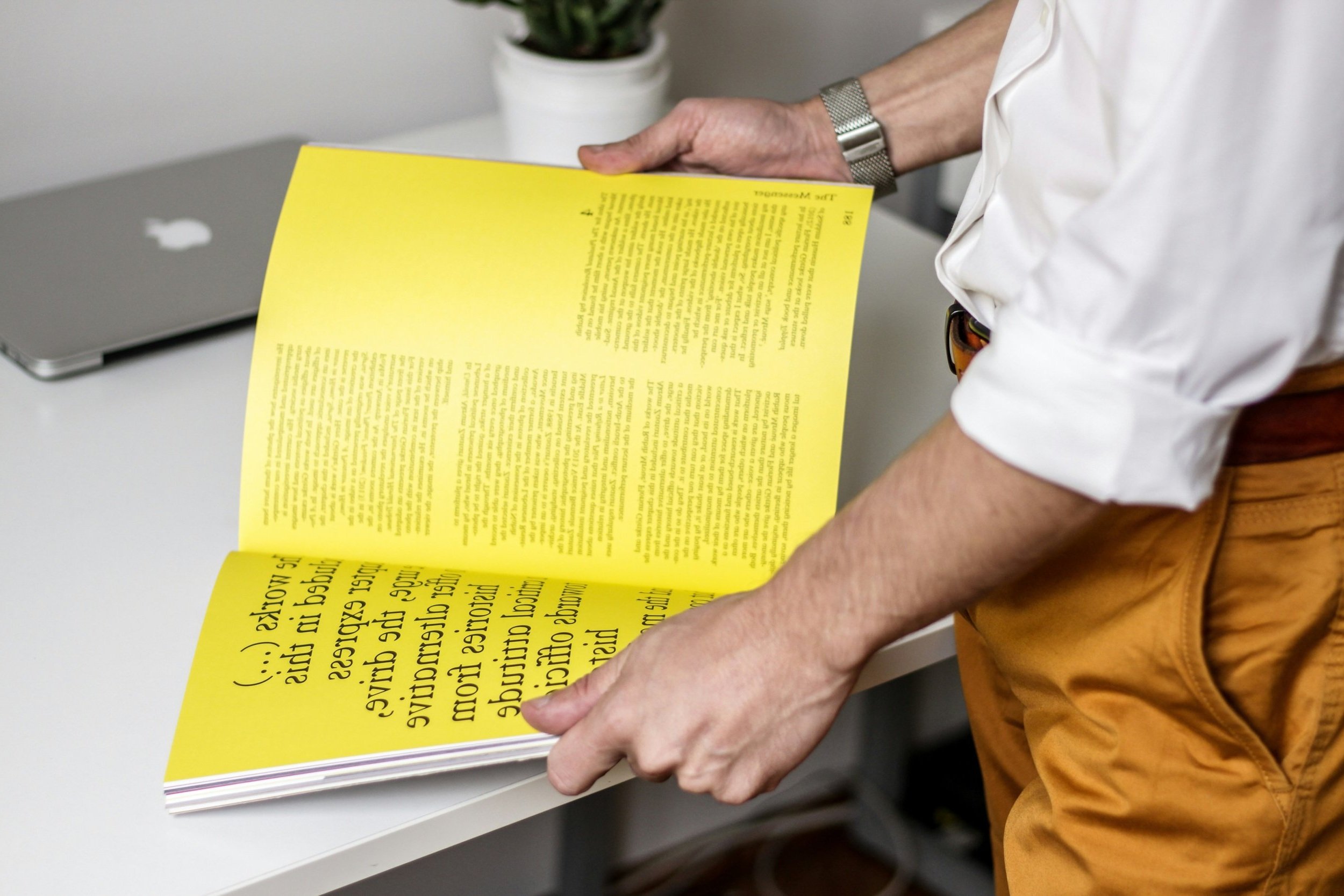 Person flipping through a bright yellow magazine or booklet at a desk, with a silver laptop and a small potted plant in the background.