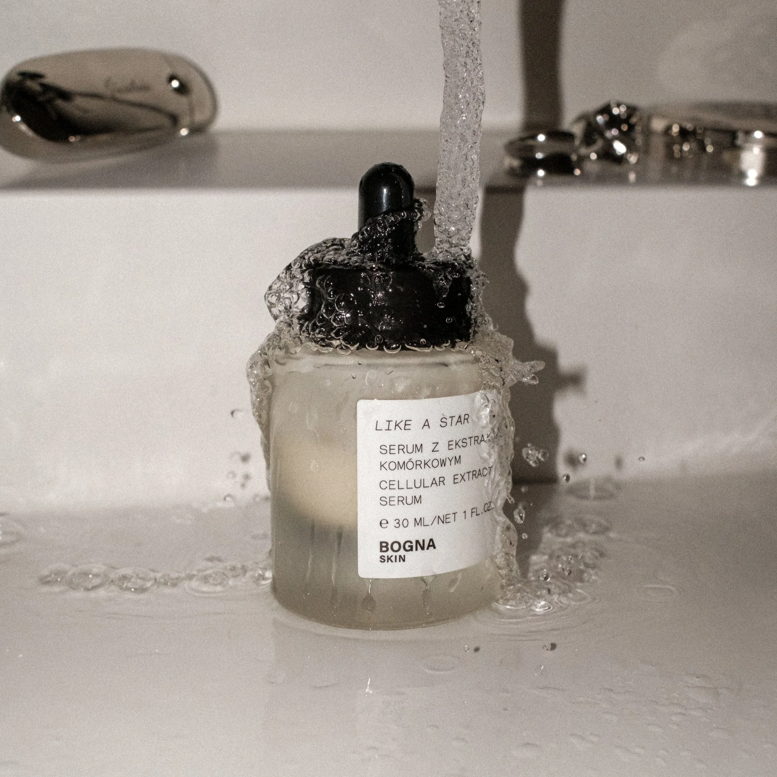 A bottle labeled 'LIKE A STAR SERUM Z EKSTRAK KOMÓRKOWYM CELLULAR EXTRACT SERUM' under running water from a faucet, with water splashing around it.
