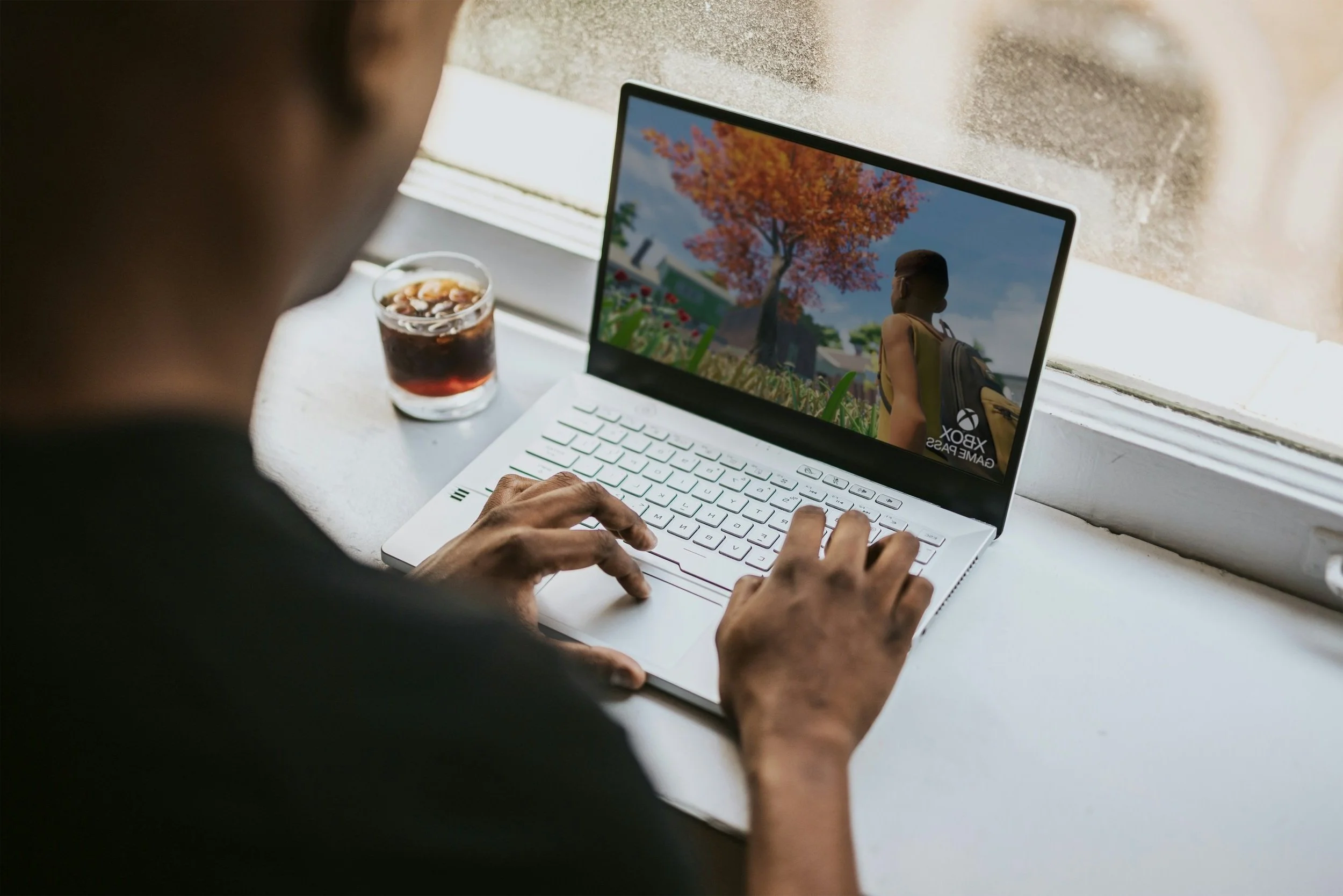 Person playing an Xbox Game Pass game on a white laptop with a cup of soda nearby, sitting by a window.