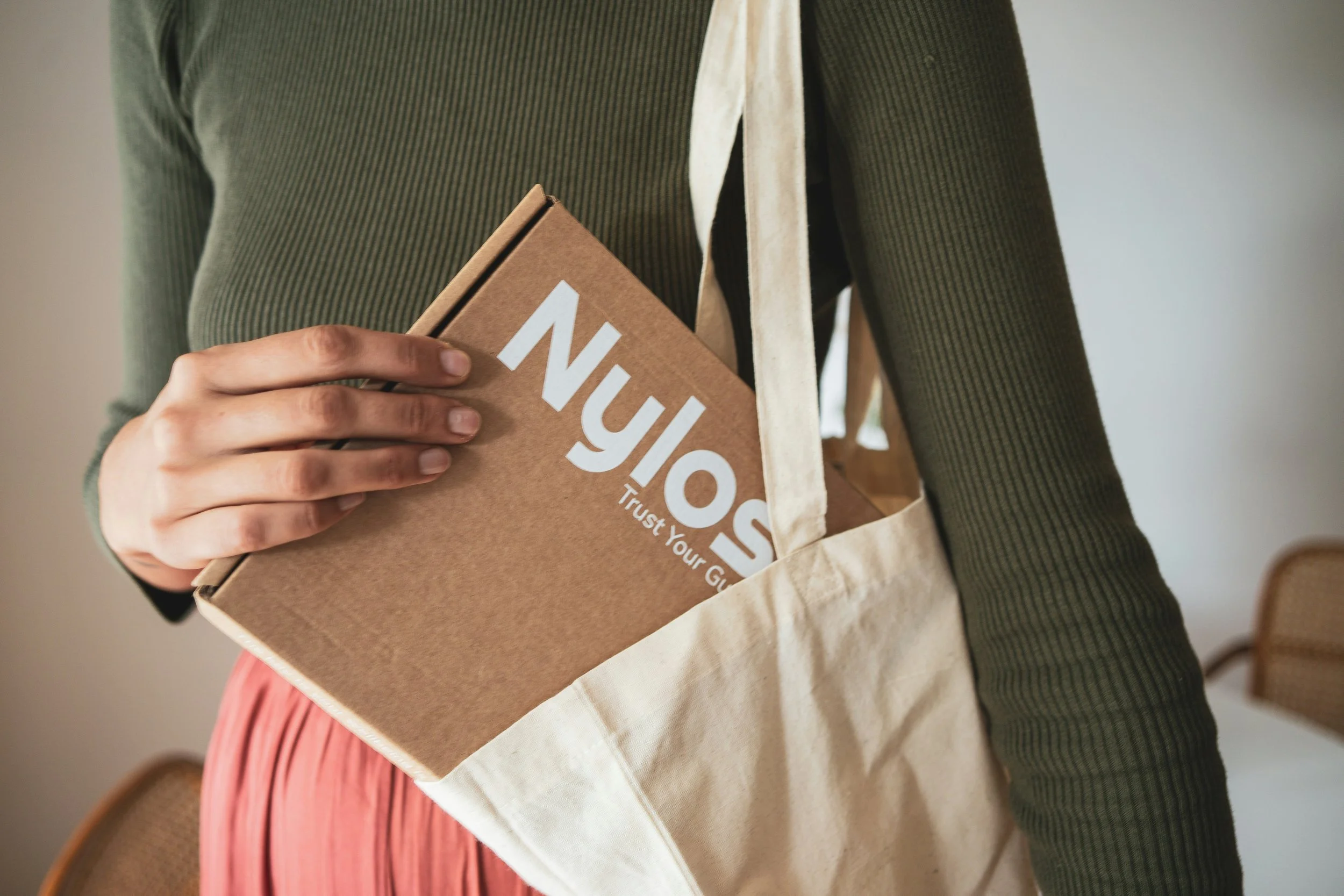 Person holding a brown cardboard box with 'Nulog' written on it, wearing a green ribbed sweater and a beige tote bag.