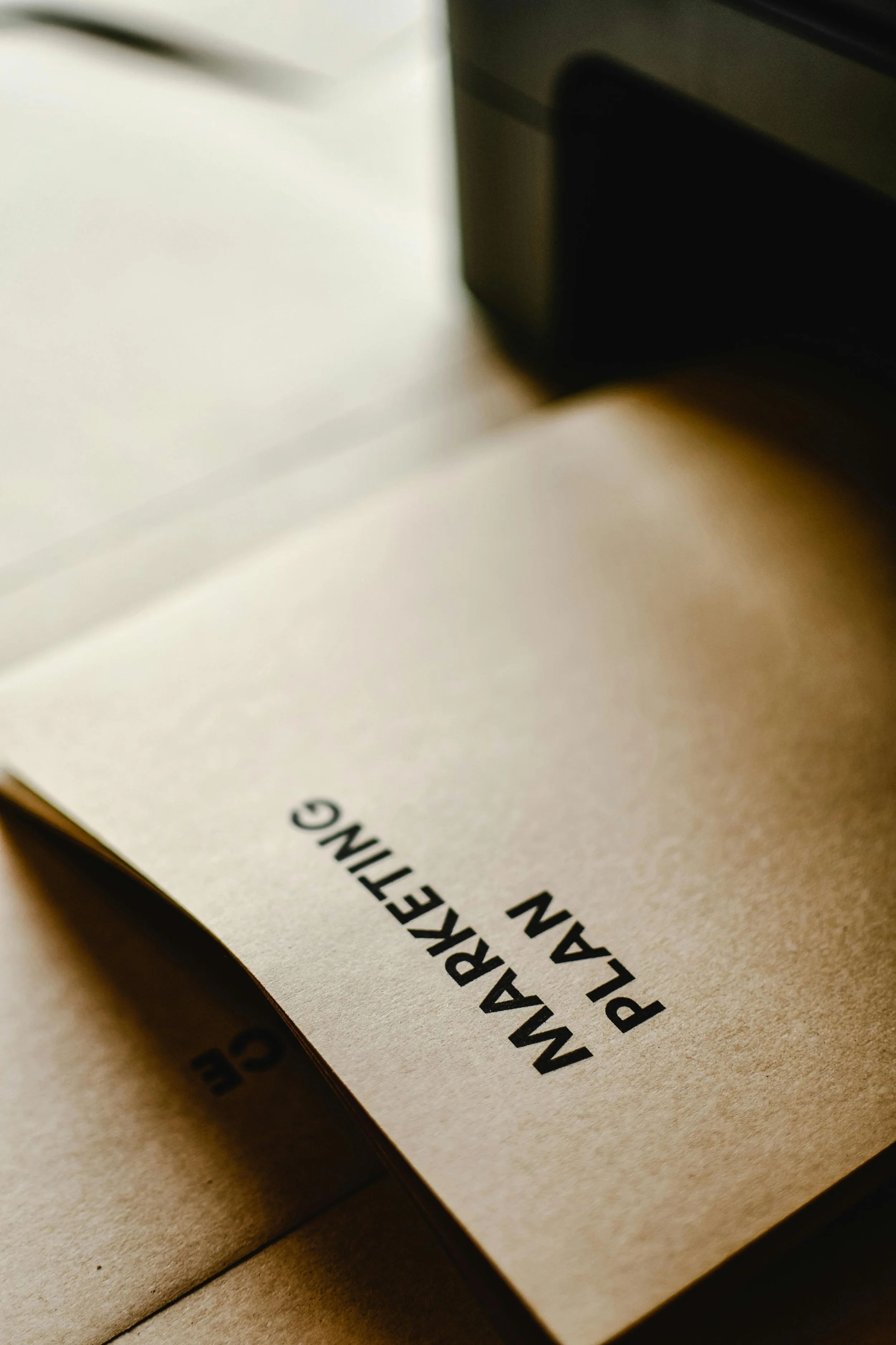 A manual with the title 'MARKETING PLAN' printed on a piece of paper, partially tucked into a brown paper envelope or folder, with a black object nearby.