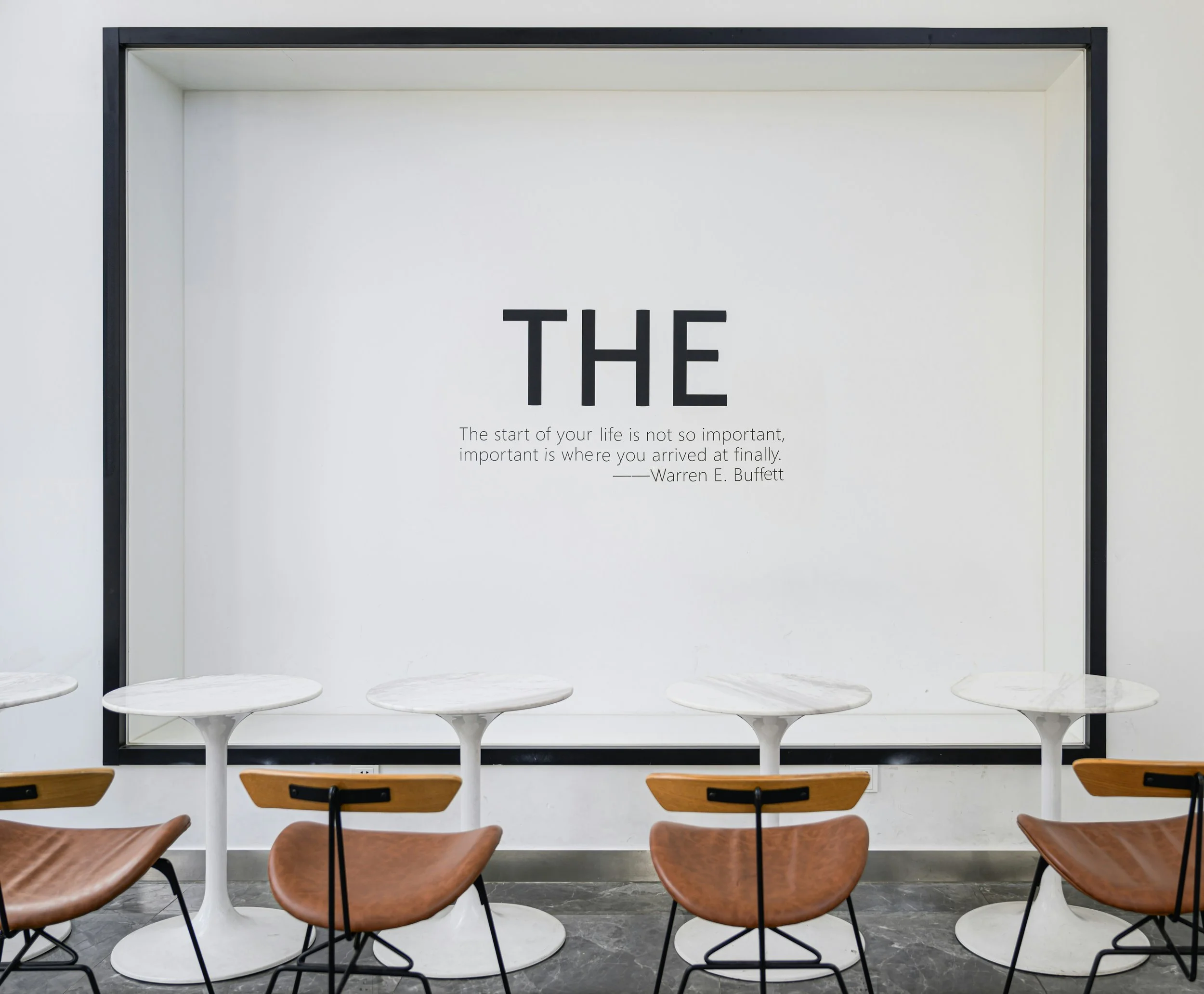 A minimalist cafe seating area with white marble tables and brown chairs, featuring a large wall quote that says 'The' and a Warren E. Buffett quote about life and its importance.