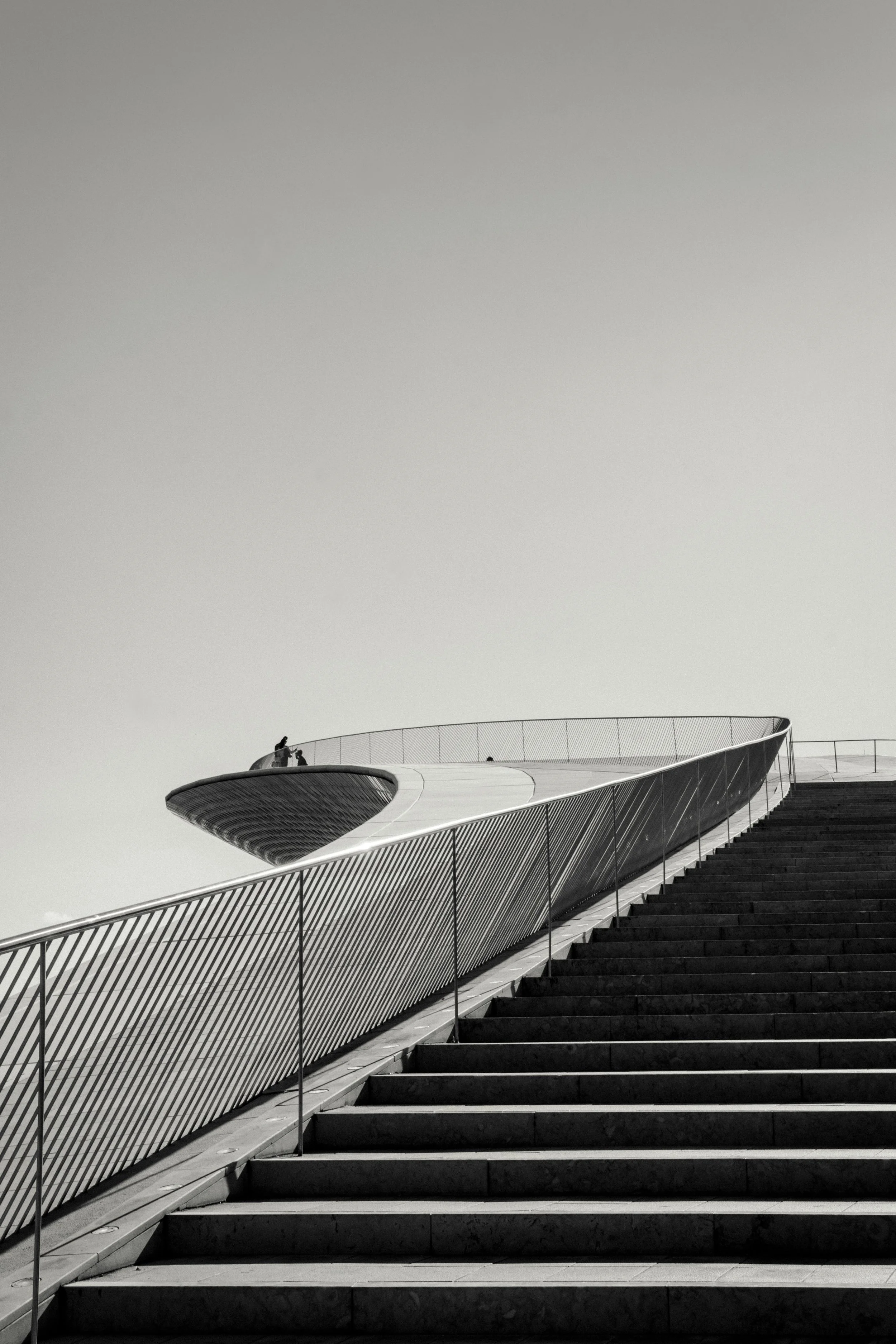 Black and white photo of a modern architectural building with a curved design, viewed from the bottom of a staircase with metal railings.