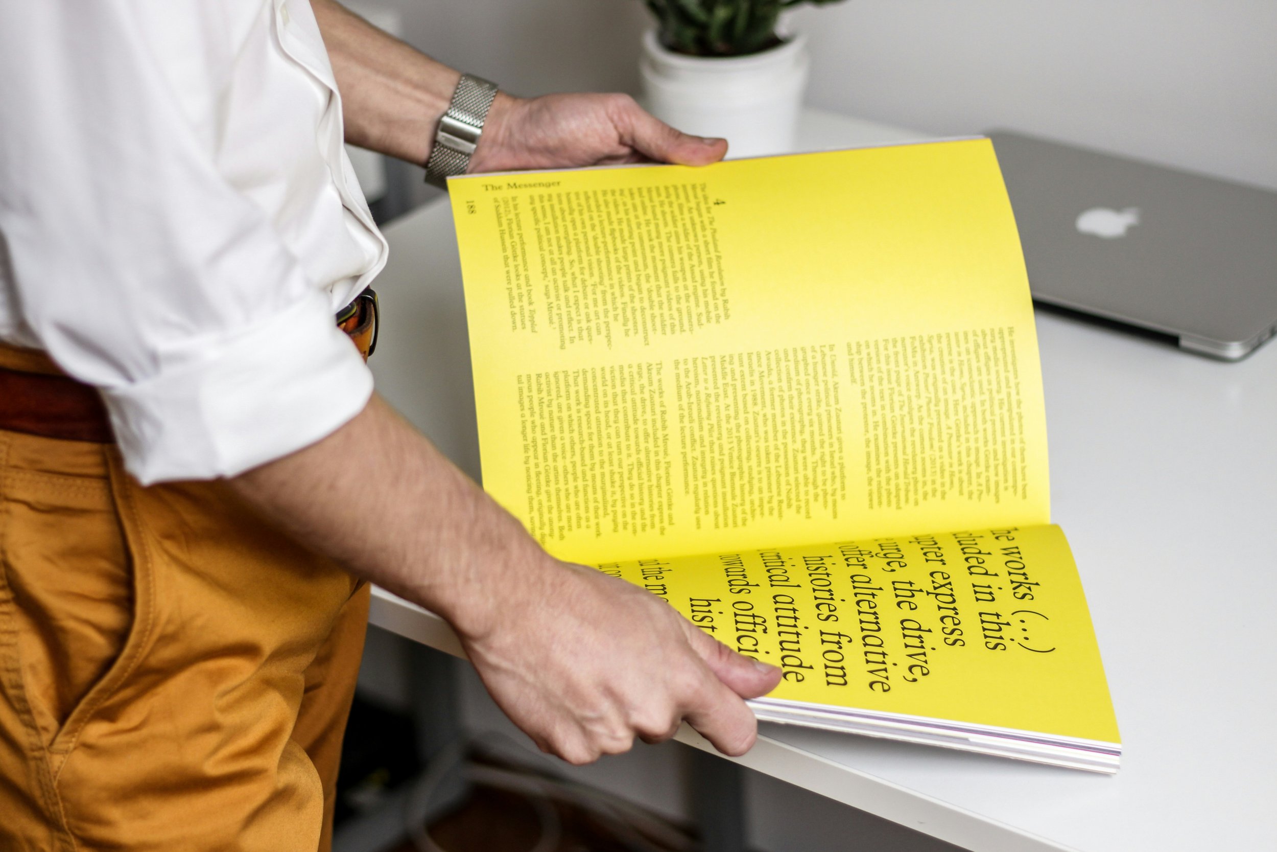 Person holding open yellow printed pamphlet or magazine with text, standing at a white table with a silver laptop and potted plant in the background.