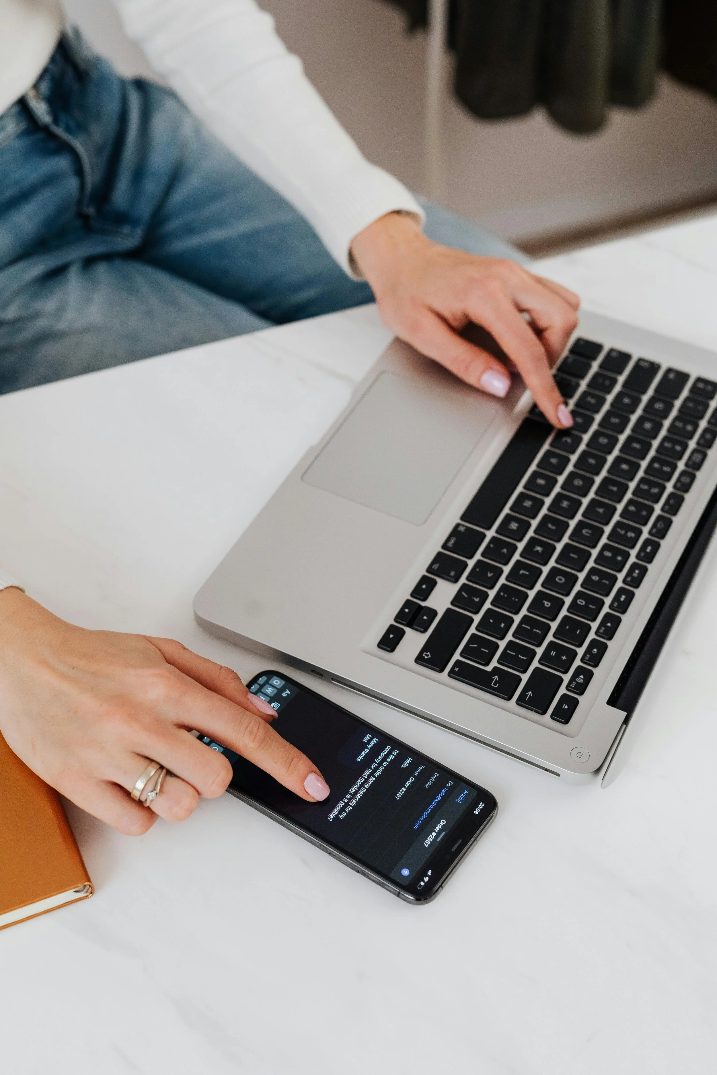 Person using a smartphone and a laptop on a white table.