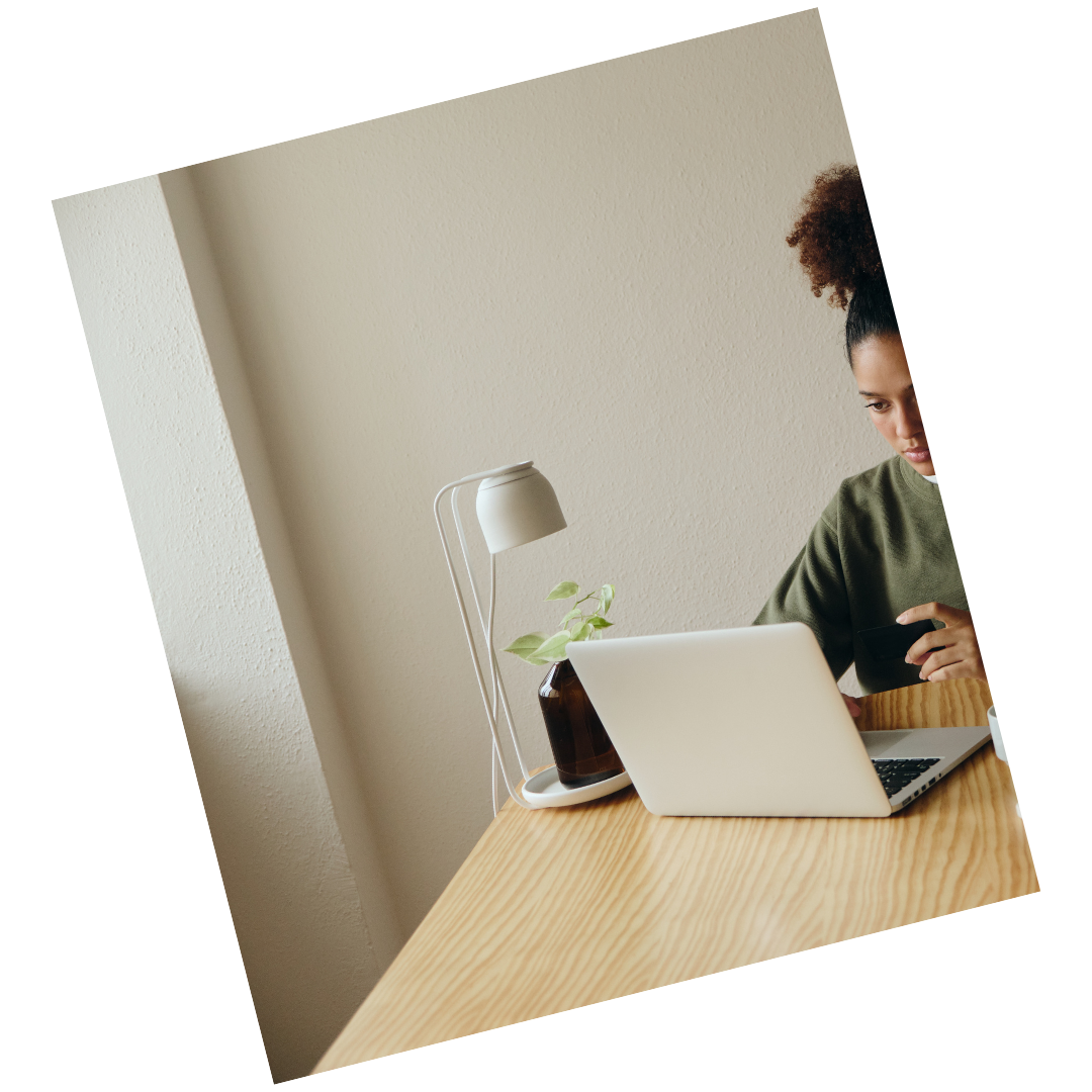 A woman with curly hair working on a laptop at a wooden desk, with a small plant and a white desk lamp nearby.