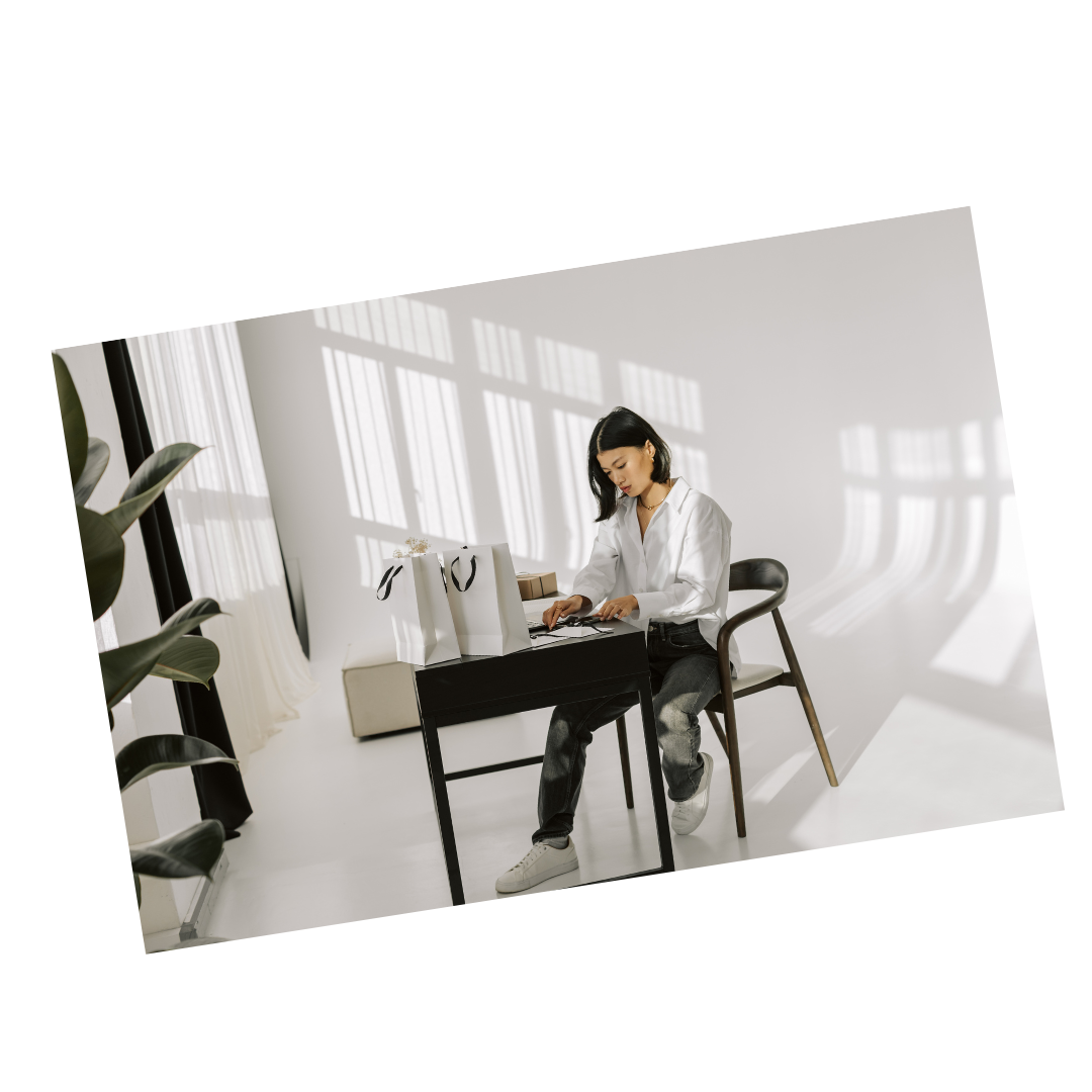 A woman with black hair, wearing a white blouse and black pants, sitting at a black desk in a well-lit room, looking down at her phone, with shopping bags on the desk and shadows of window frames cast on the white wall behind her.