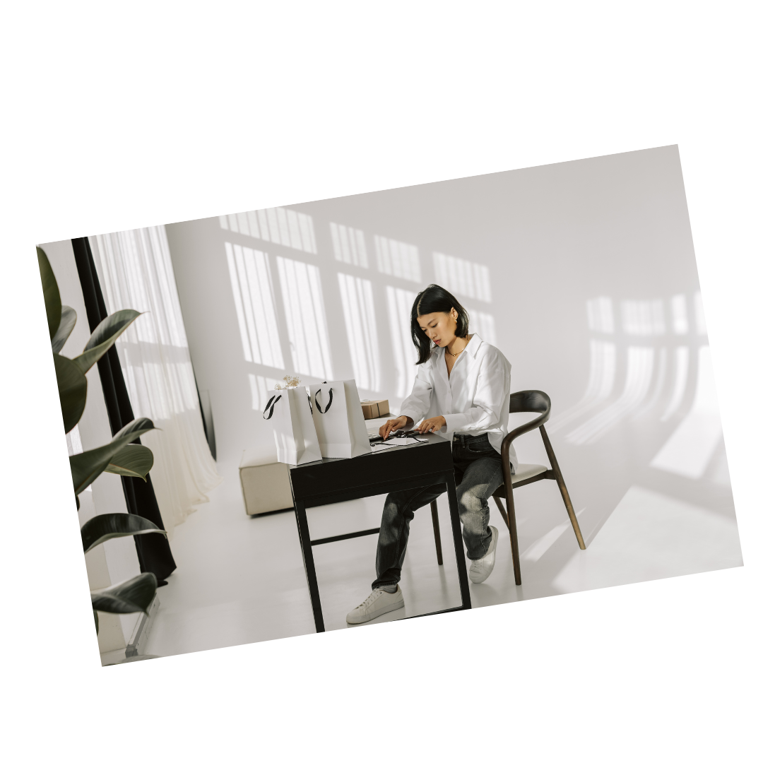 A woman sitting at a black desk with shopping bags and boxes, working on a laptop in a bright room with large windows and natural light.