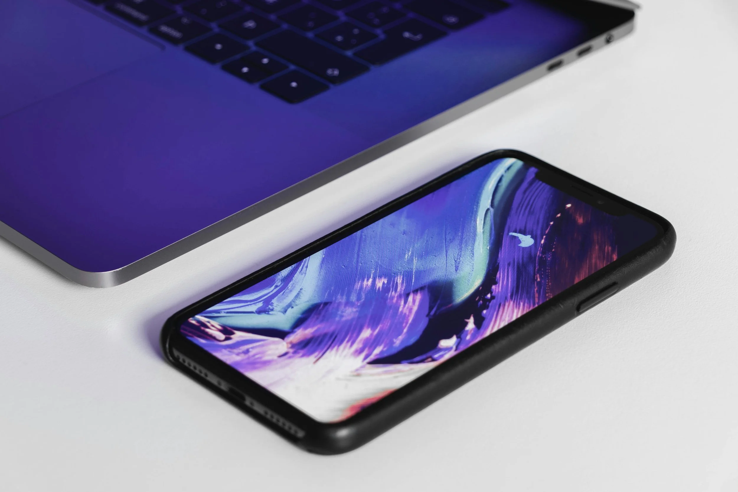 An iPhone with a colorful abstract display placed next to a silver laptop with a black keyboard on a white surface.