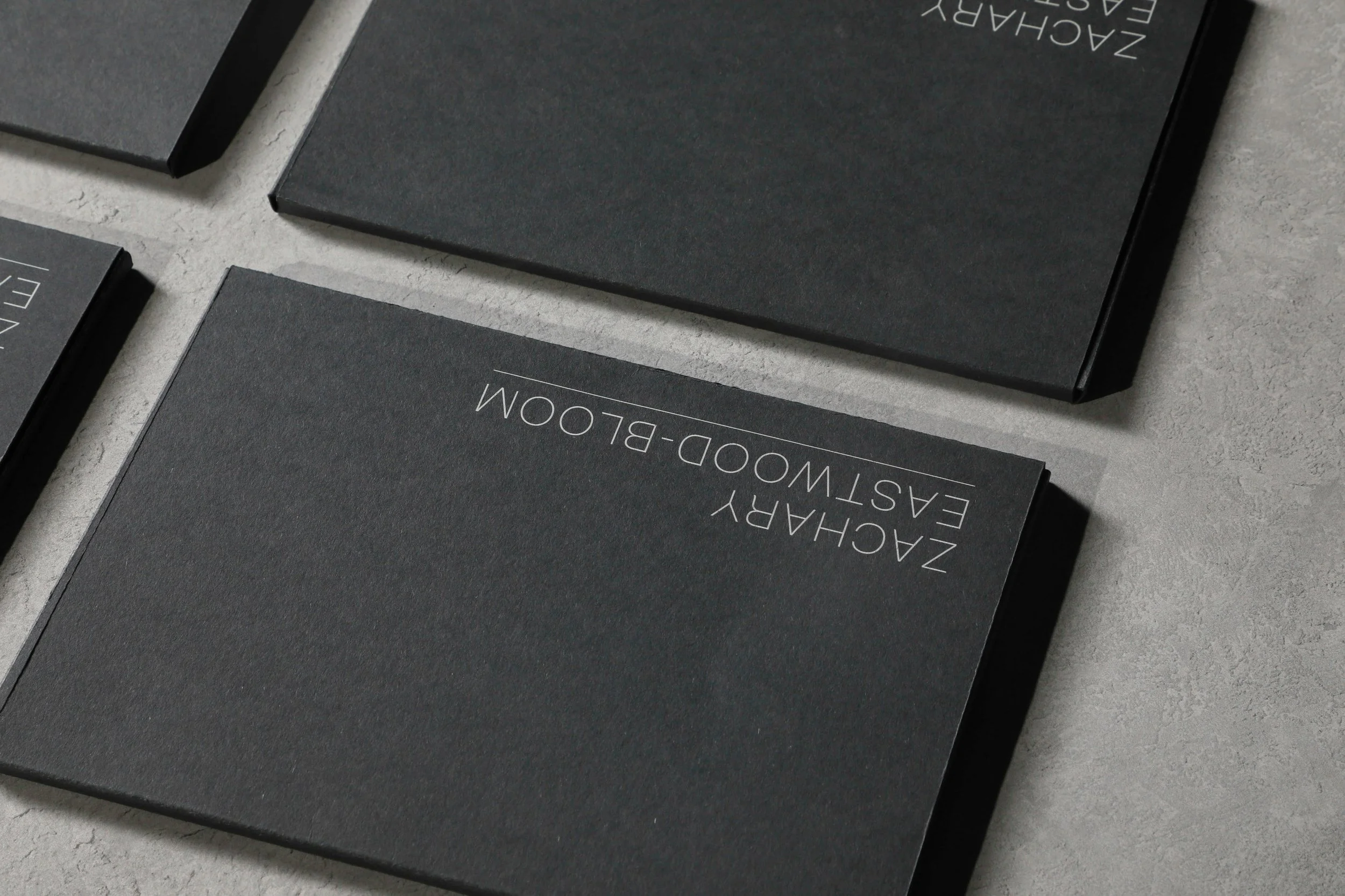 Black packaging boxes with white text reading 'Zachary Eastwood-Bom' arranged on a textured grey surface.