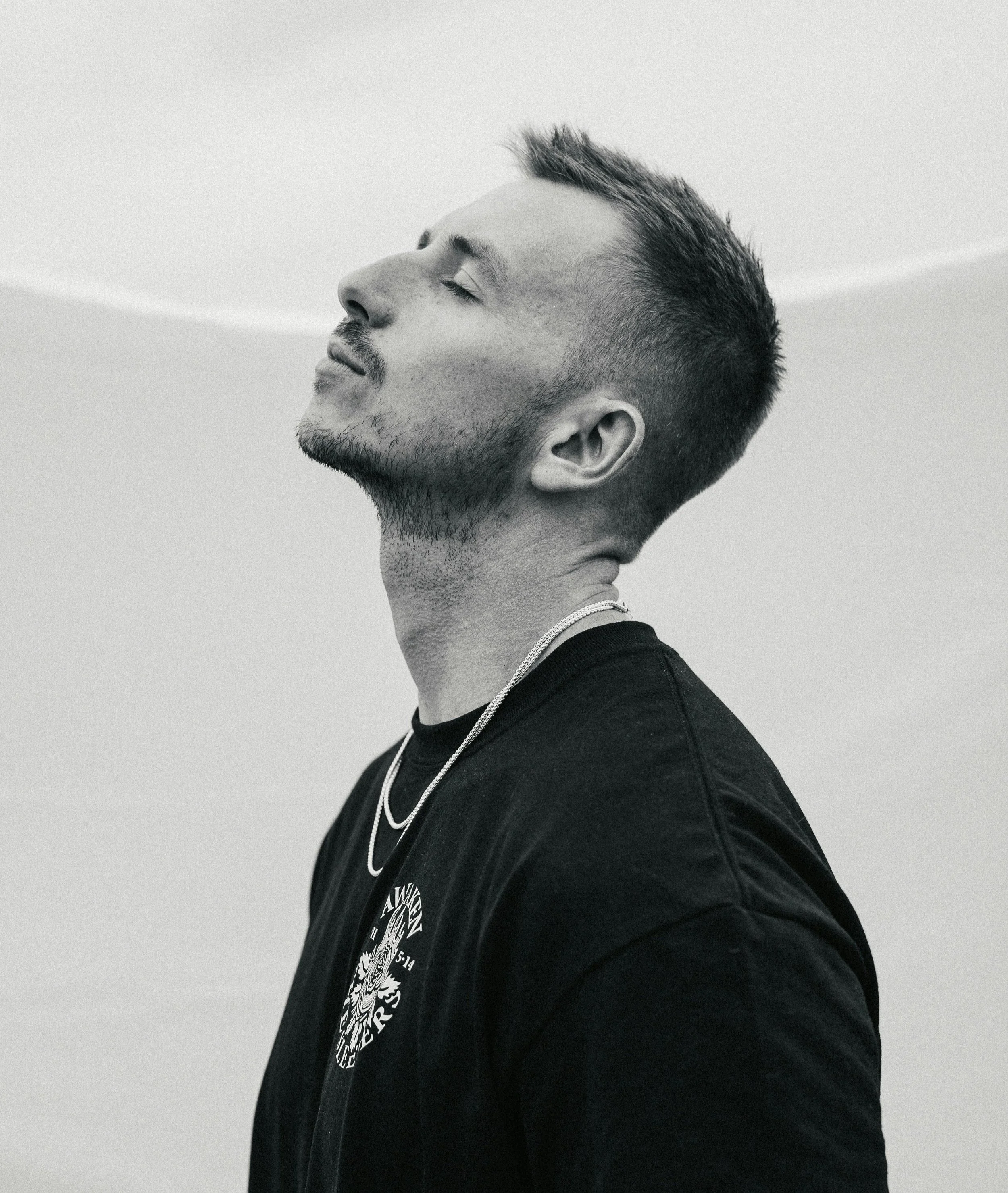 A young man with short hair and a beard, wearing a black t-shirt and chain necklaces, is seen with his eyes closed and head tilted back, enjoying a moment of relaxation.
