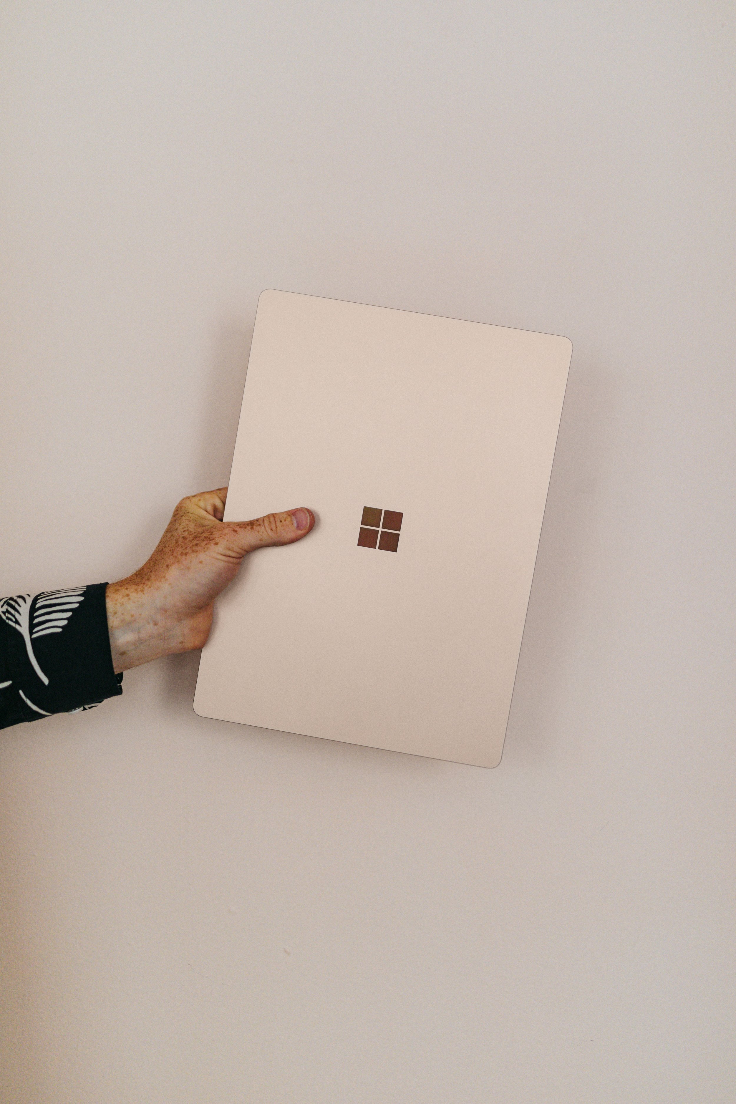 A hand holding a white Microsoft Surface device with the Microsoft logo on the back, positioned against a plain white wall.