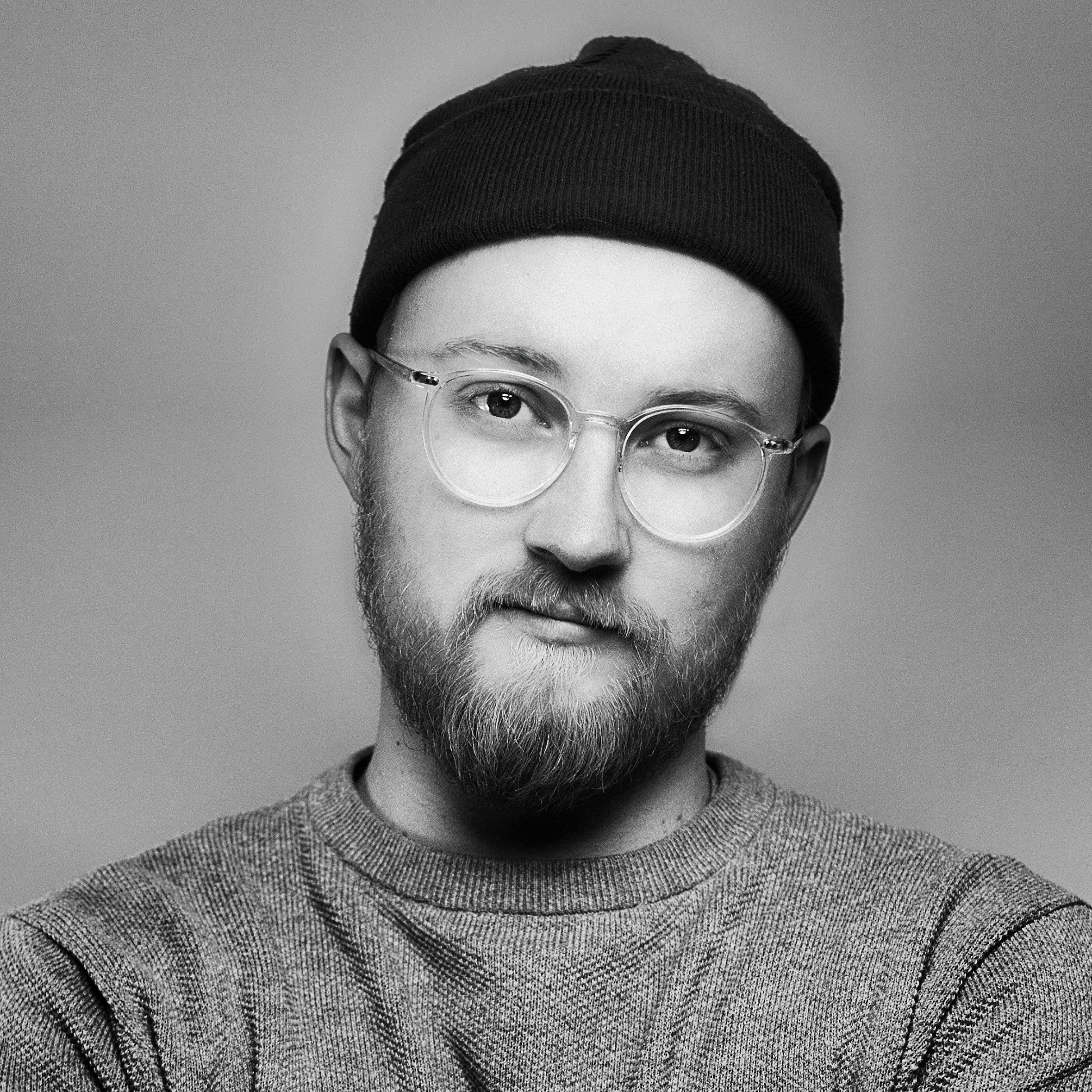 Black and white portrait of a young man with a beard, round glasses, a beanie, and a casual sweater, looking at the camera with a slight smile.