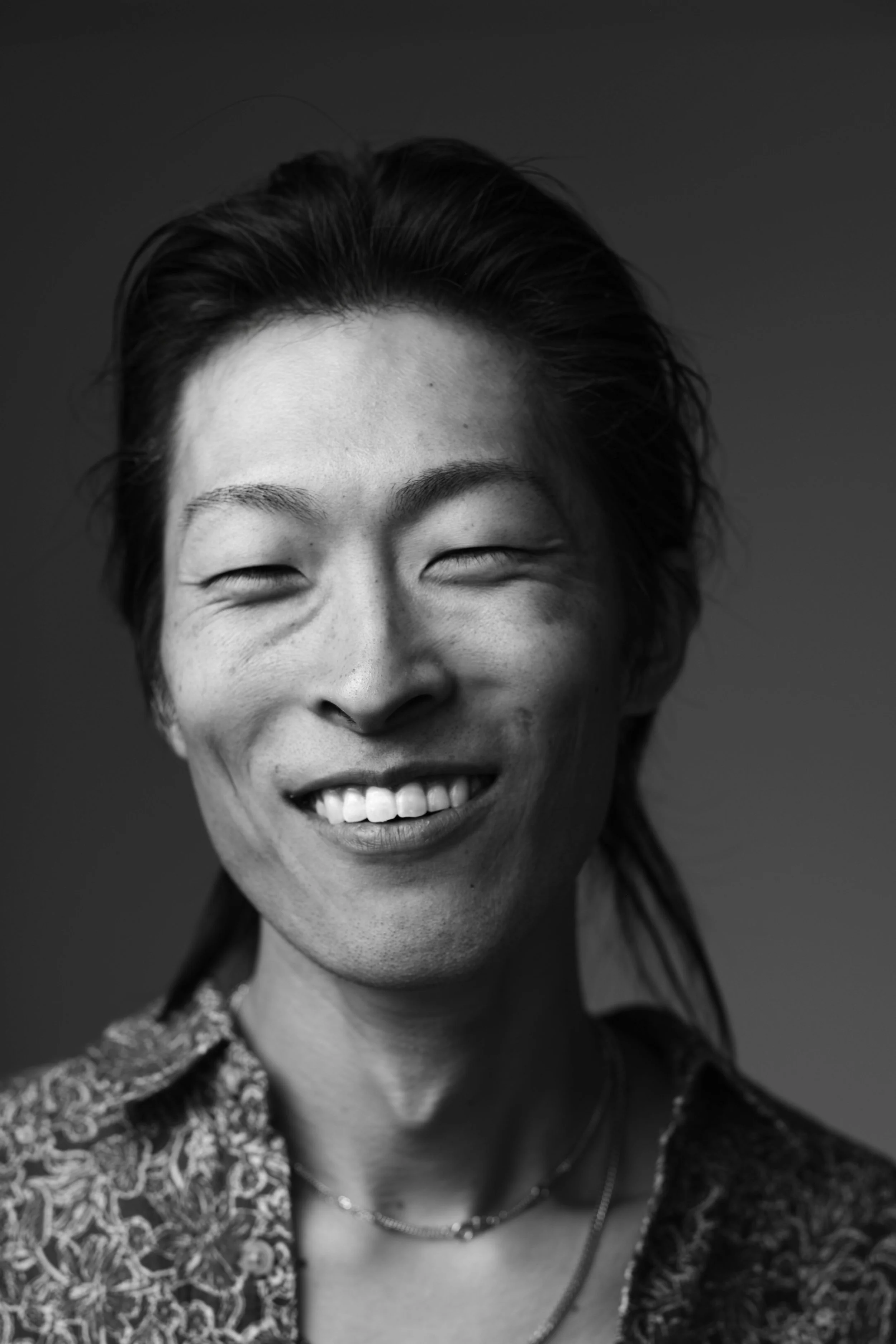 Black and white portrait of a woman smiling with eyes closed, wearing a patterned jacket and layered necklaces.