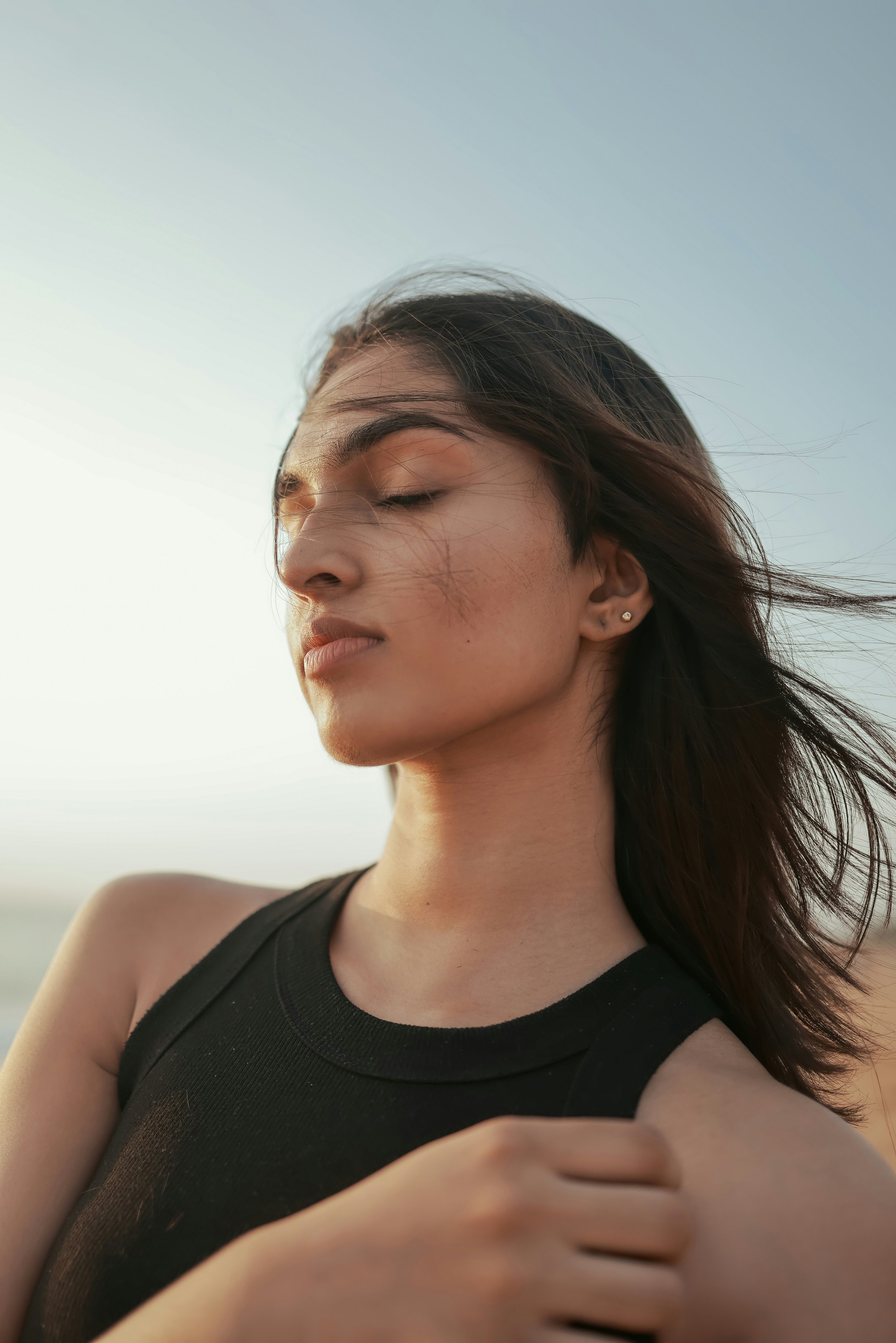 A woman with long dark hair and earrings wearing a black sleeveless top, standing outside with her eyes closed, during sunset.