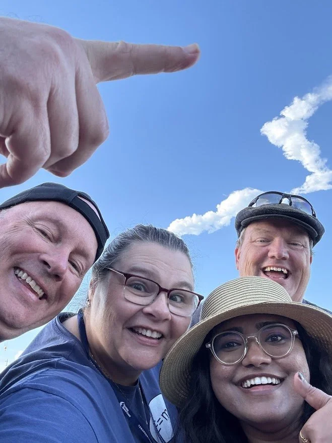Four people taking a selfie with the rocket trail in the background.  The person on the left is pointing at the trail.