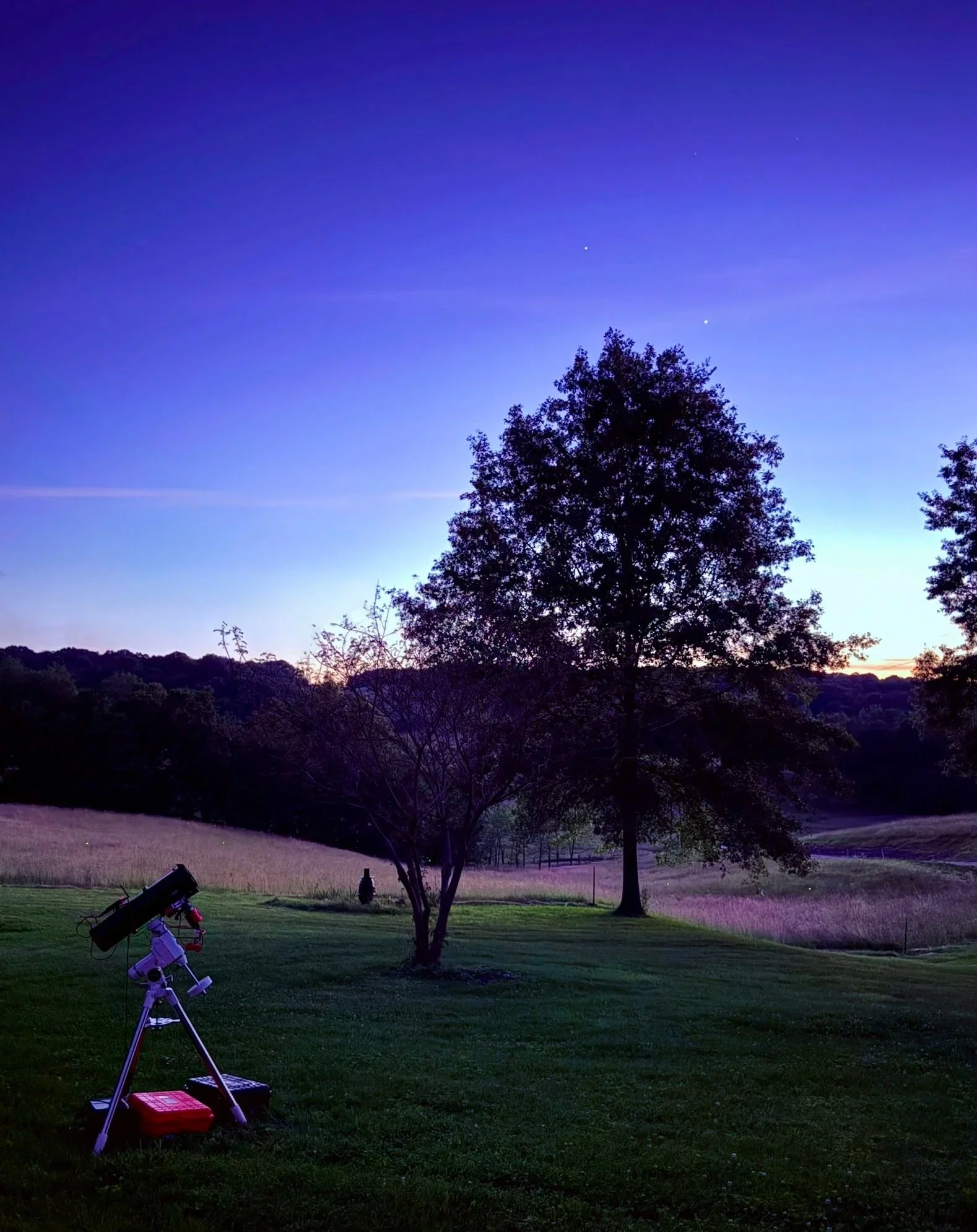 A telescope at dusk in a field with a tree with two bright objects (Jupiter & Venus) in the sky.