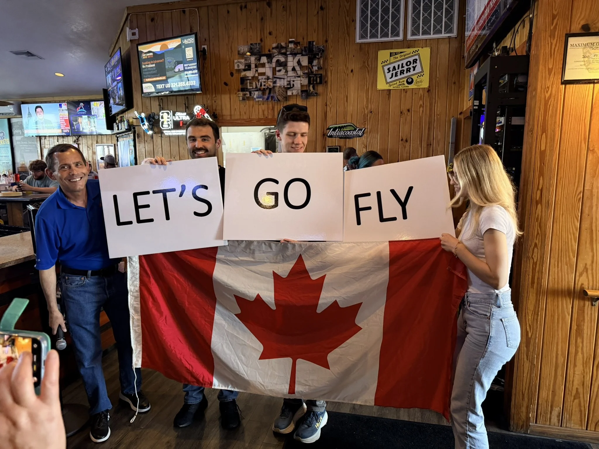 People in a bar holding a Canadian Flag along with signs that say "Let's Go Fly!"