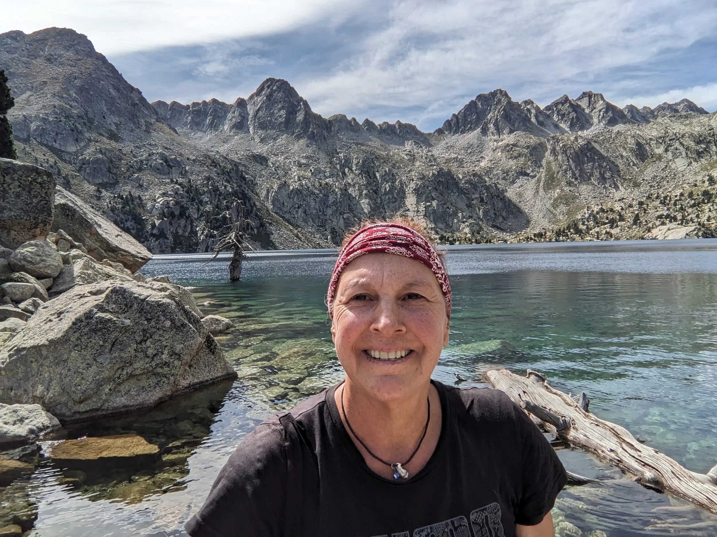 Eine lachende Frau mit roten Haarband am Ufer eines klaren Sees inmitten von Bergen und Felsen, die im Hintergrund majestätische Berge zeigen.