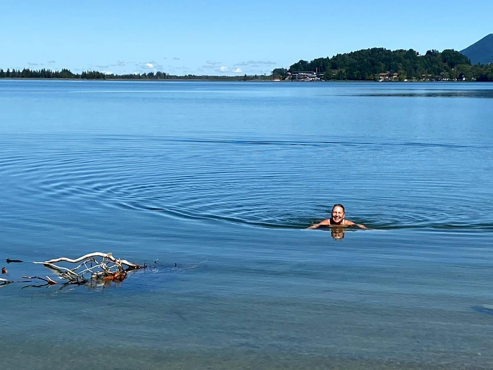 Eine Frau schwimmt lachend im ruhigen See bei Sonnenschein, mit Wald und Berge im Hintergrund.