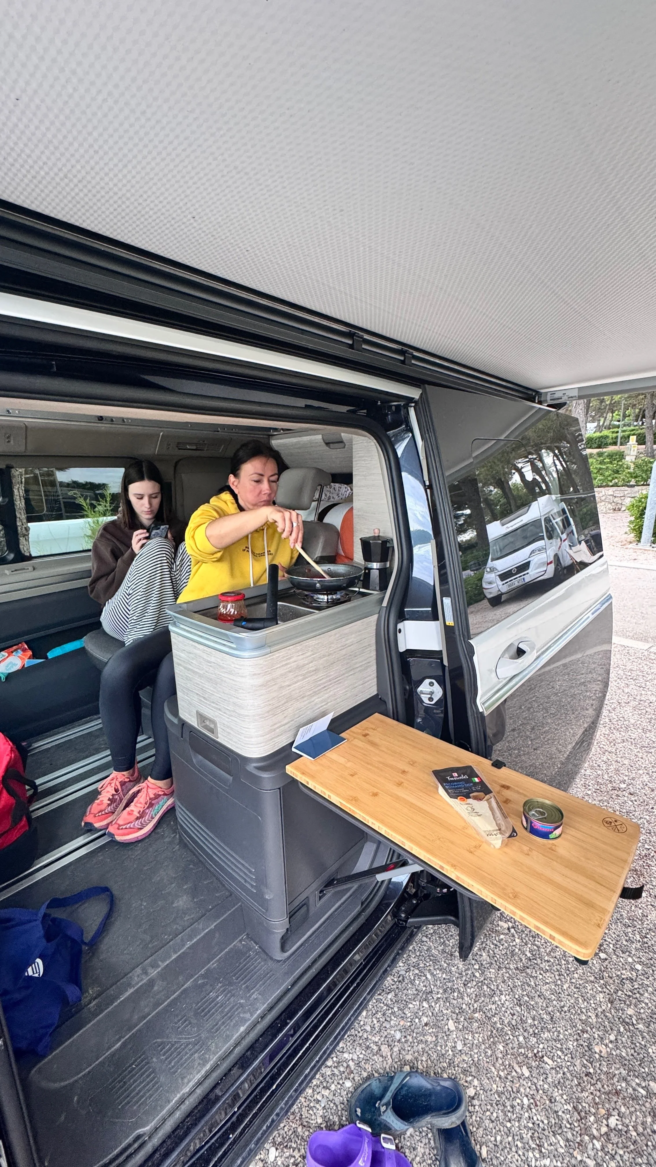 A woman cooking on a portable stove inside a camper van parked outdoors, with a girl sitting behind her using a smartphone.