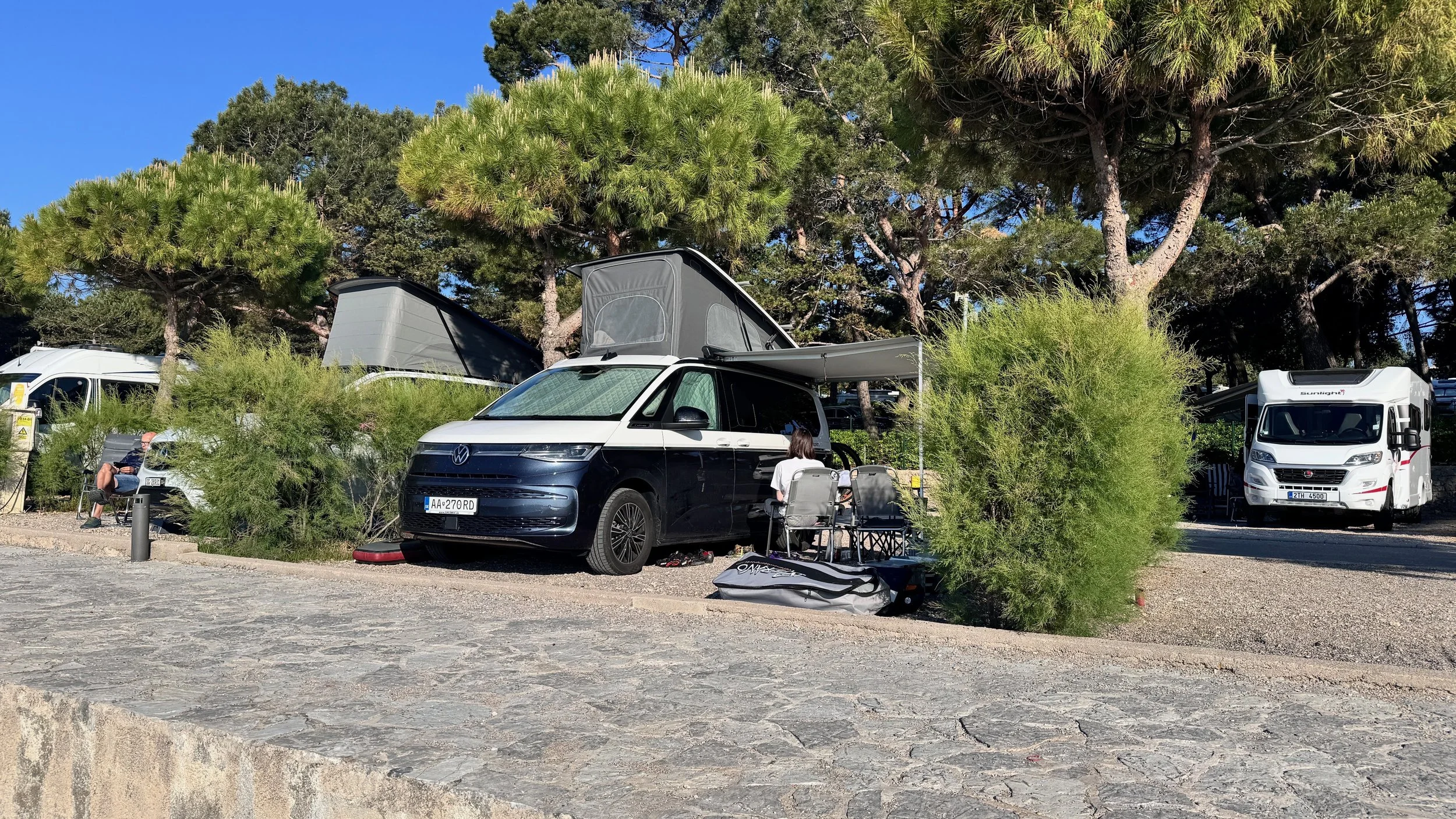 A camper van with a pop-up roof parked among trees and bushes in a campsite, with people relaxing and camping equipment around.